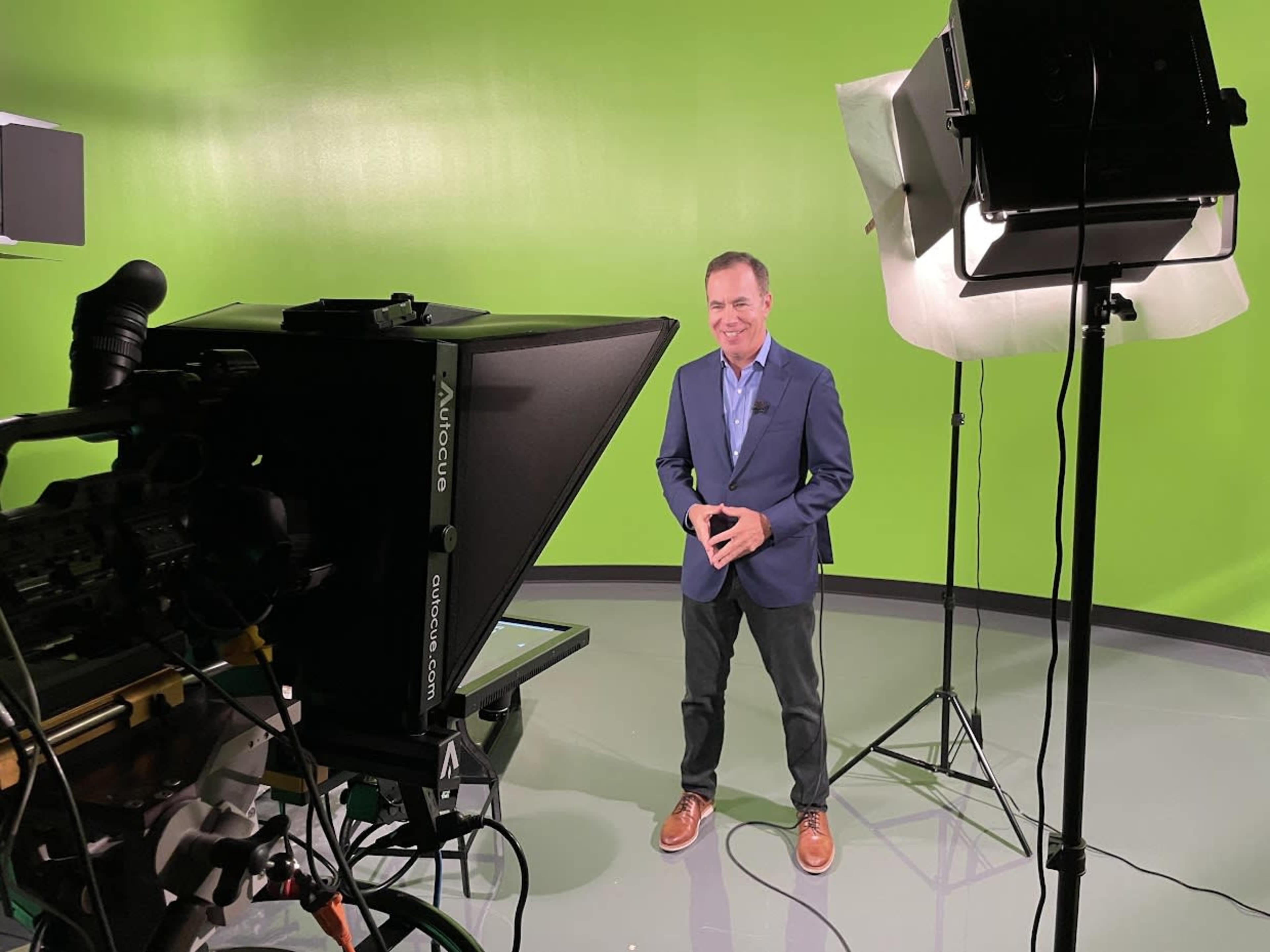 A man stands in front of a green screen while being filmed by a camera on a tripod.