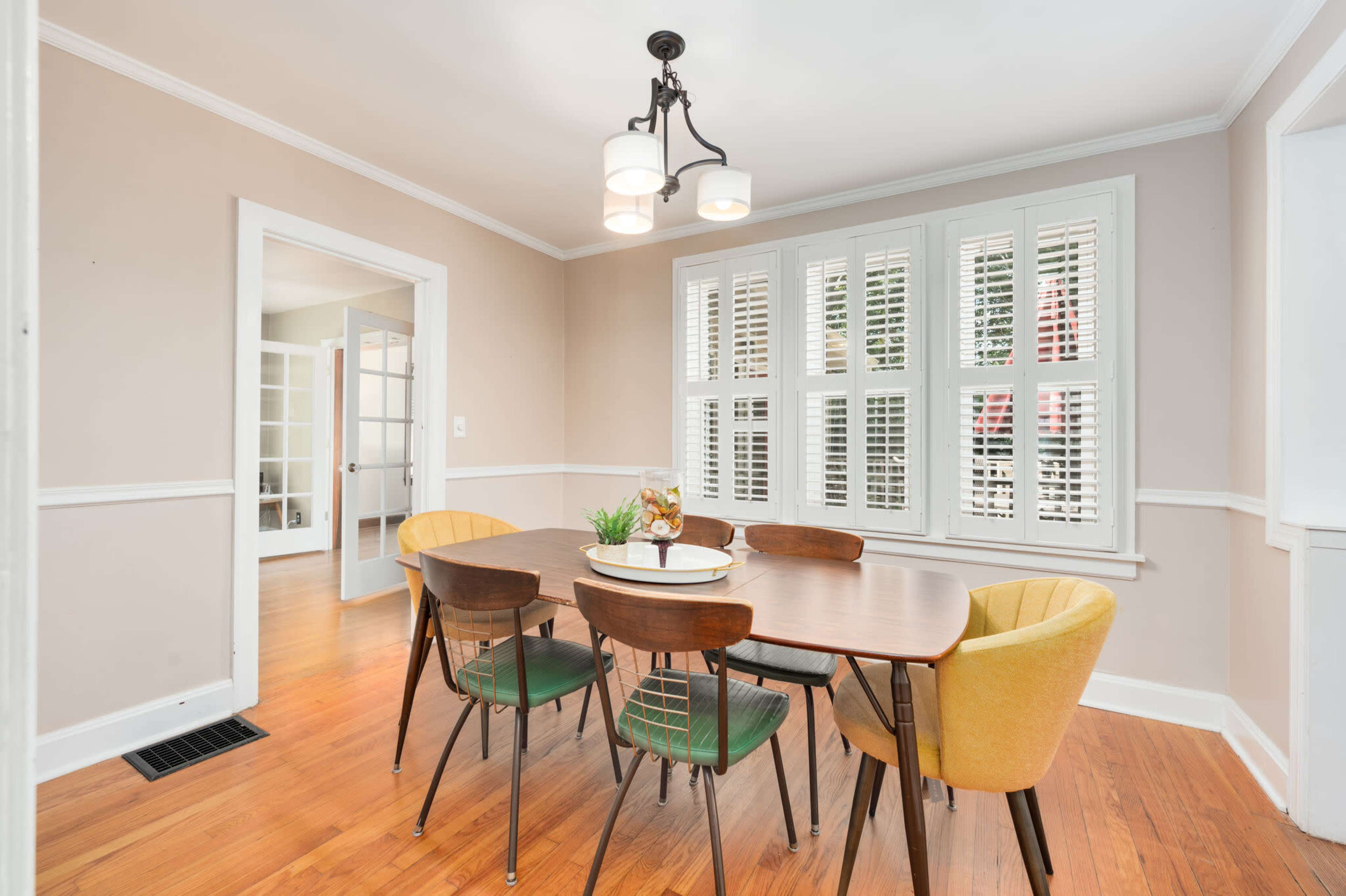 A dining room features a round wooden table surrounded by colorful chairs and a chandelier above, with windows adorned by shutters.