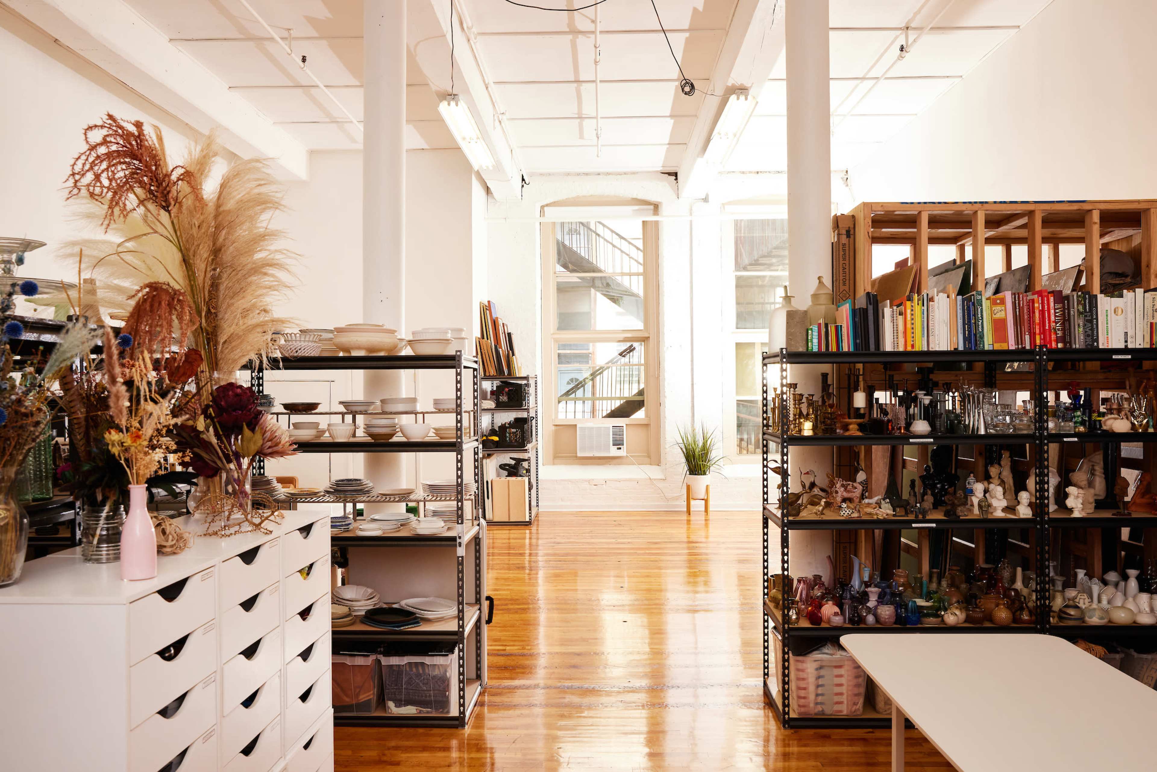 The image shows a spacious studio with shelves filled with various items, including books, dishes, and decorative objects, and large windows visible in the background.