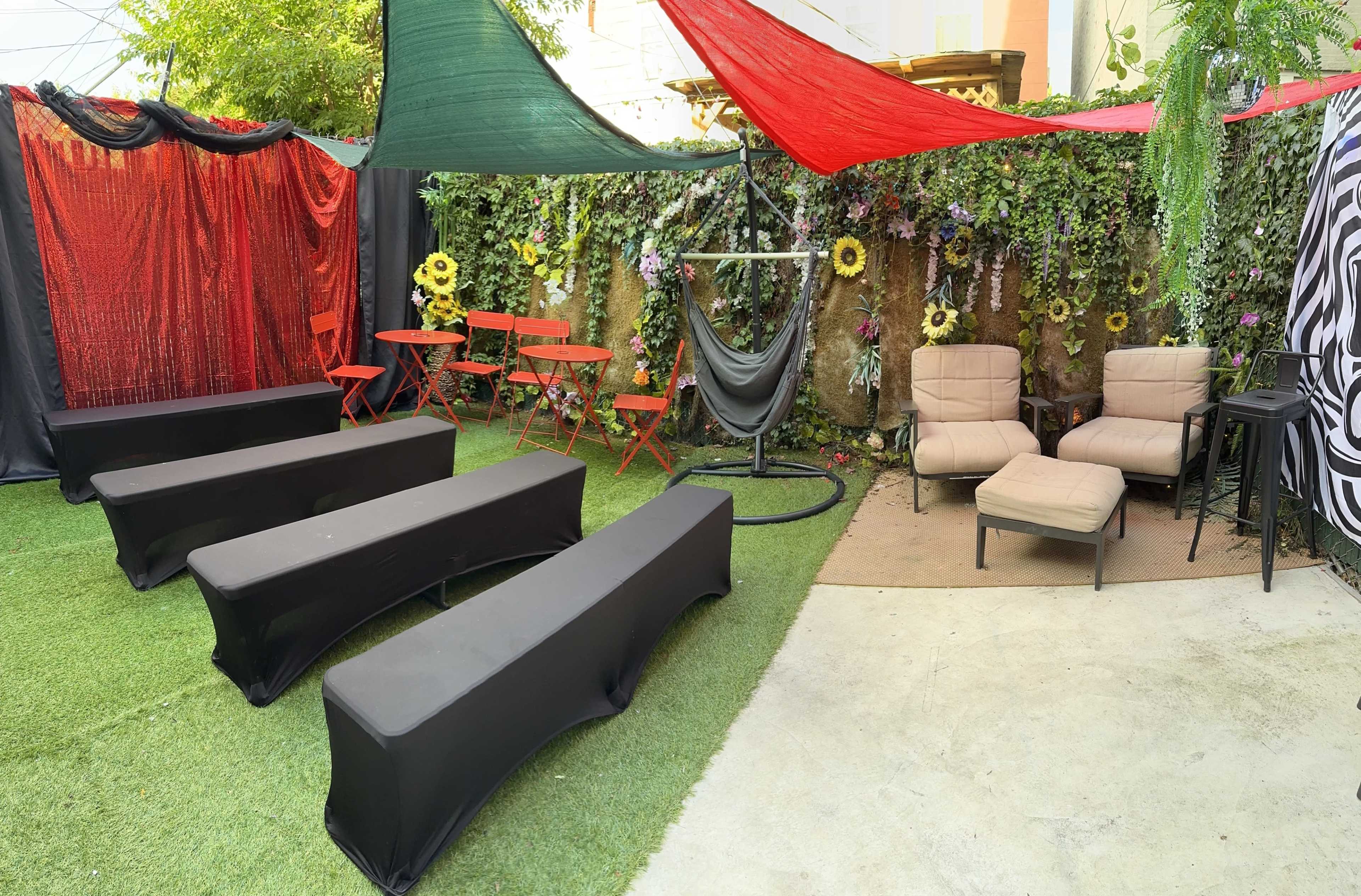A colorful outdoor space featuring black seating, red and green canopies, and floral decorations, with two armchairs and a hammock in a corner.