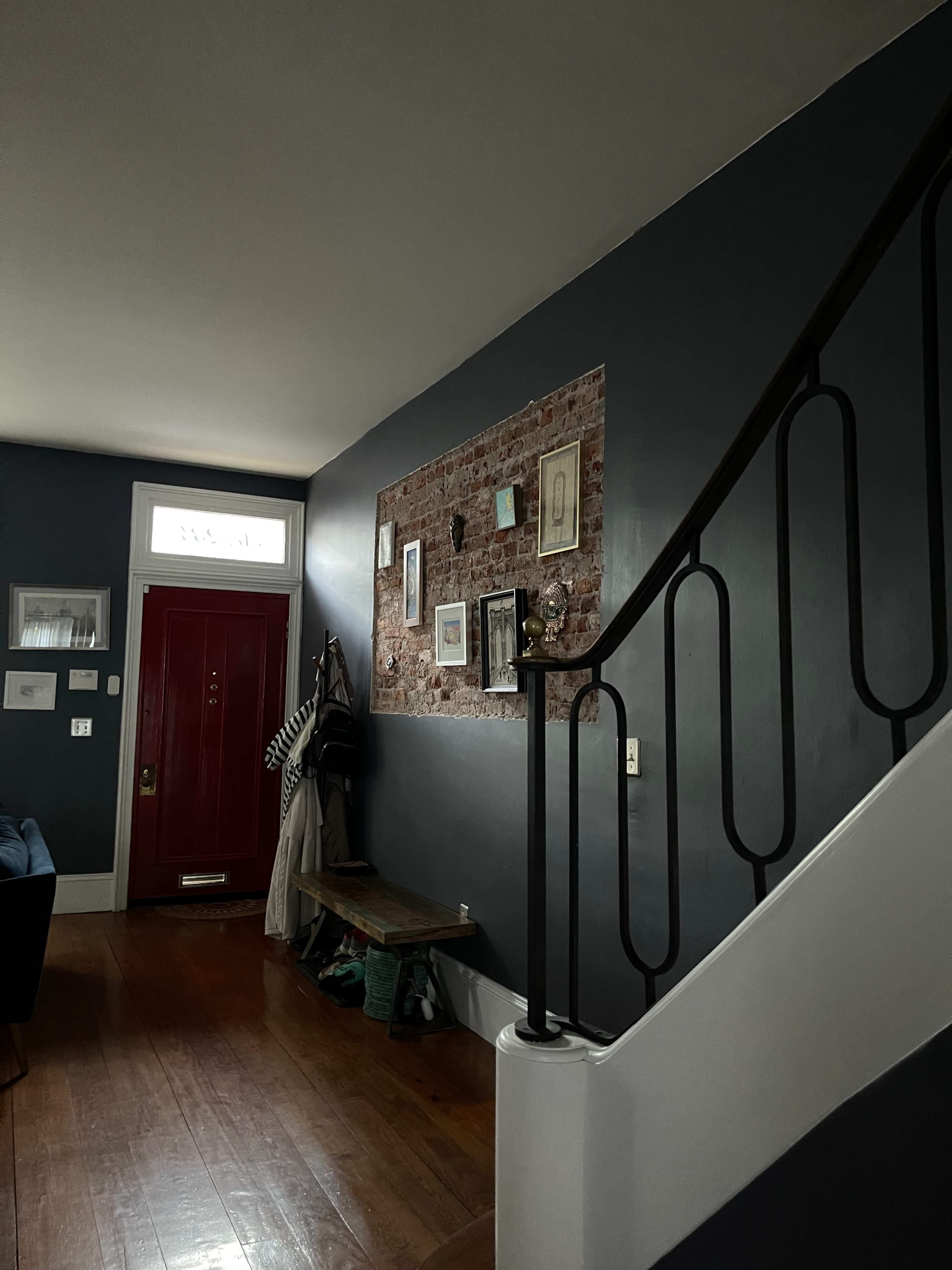 A red door is visible at the front of a hallway featuring a brick accent wall adorned with various framed pictures, alongside a staircase and a coat rack.