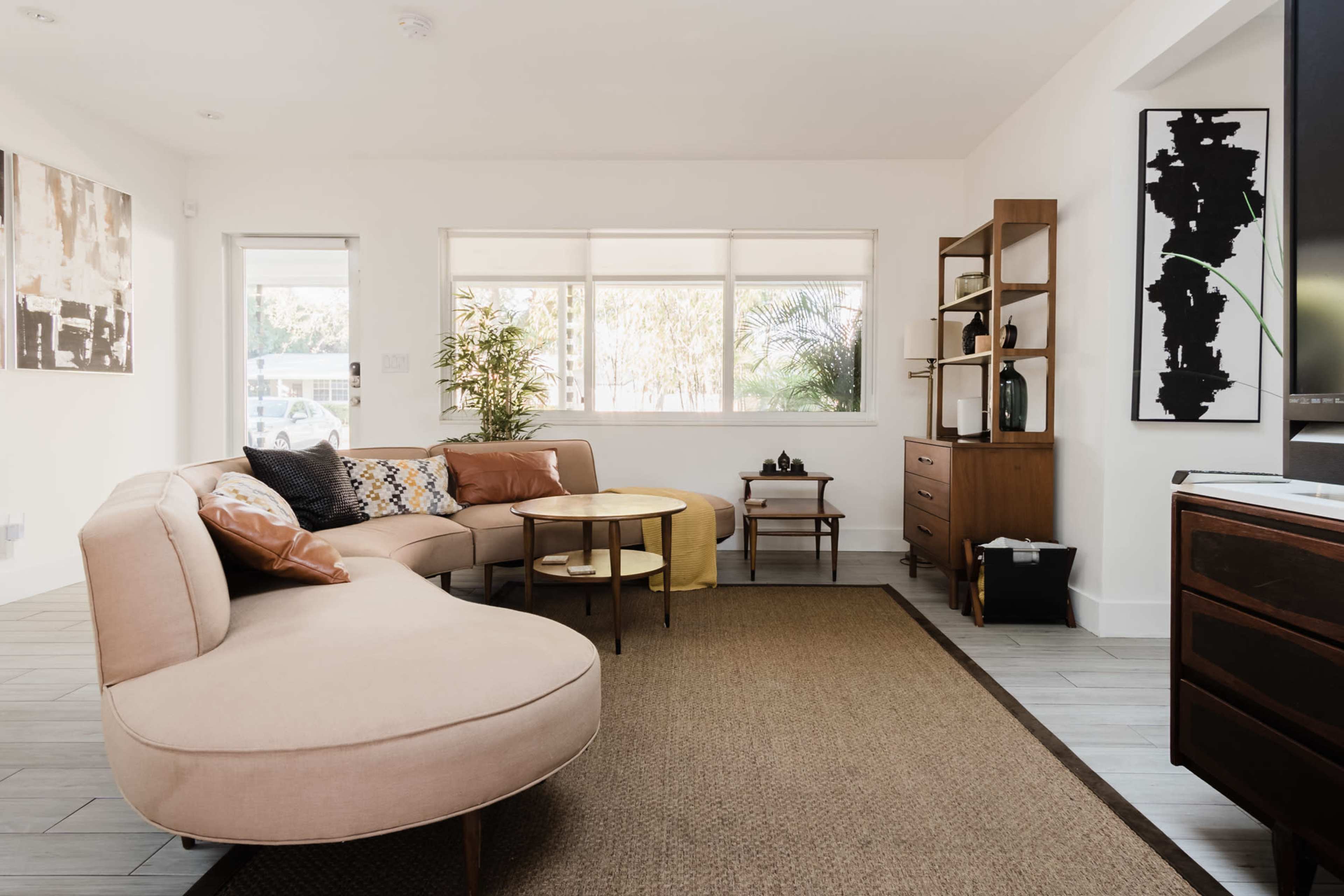 A modern living room featuring a curved beige sofa, two coffee tables, and a wooden shelving unit against light-colored walls and large windows.