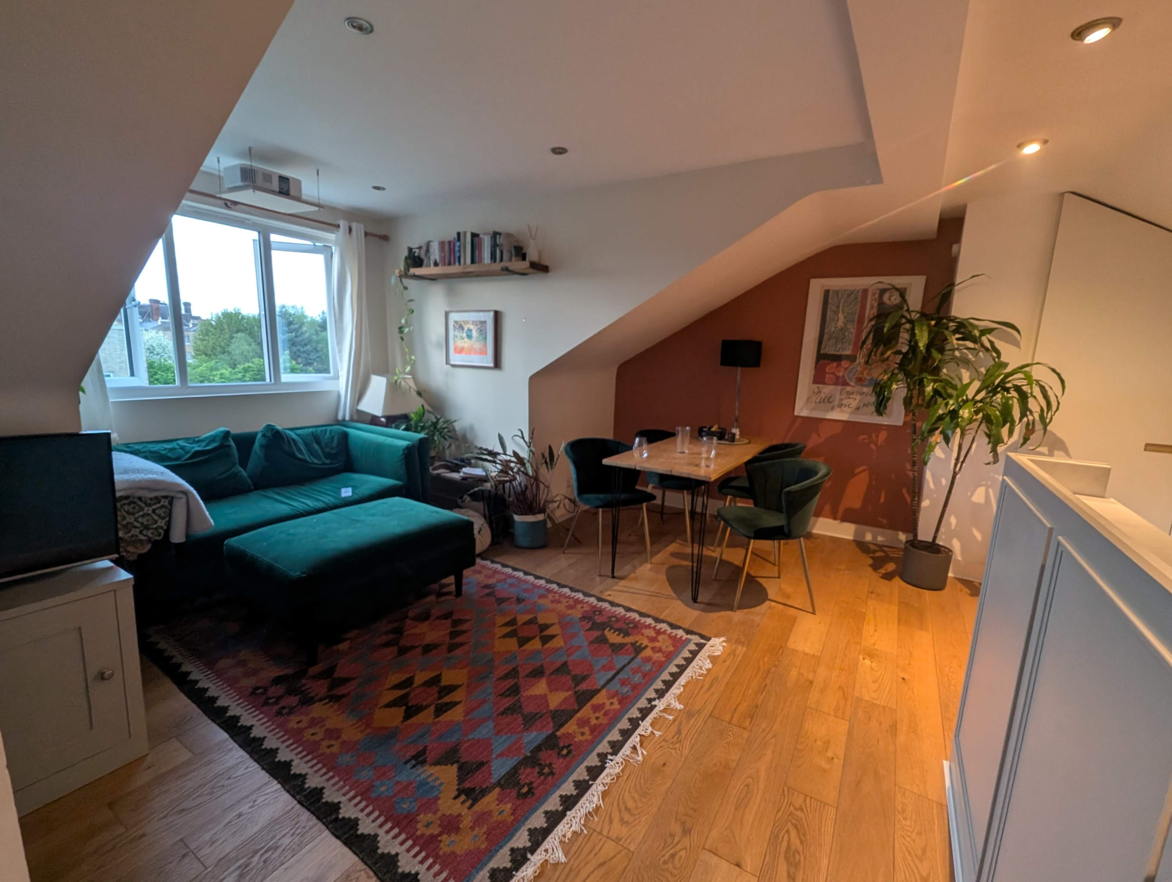 The image shows a cozy living room area with a green sectional sofa, a round dining table with chairs, and large windows providing natural light, set in a modern attic space.