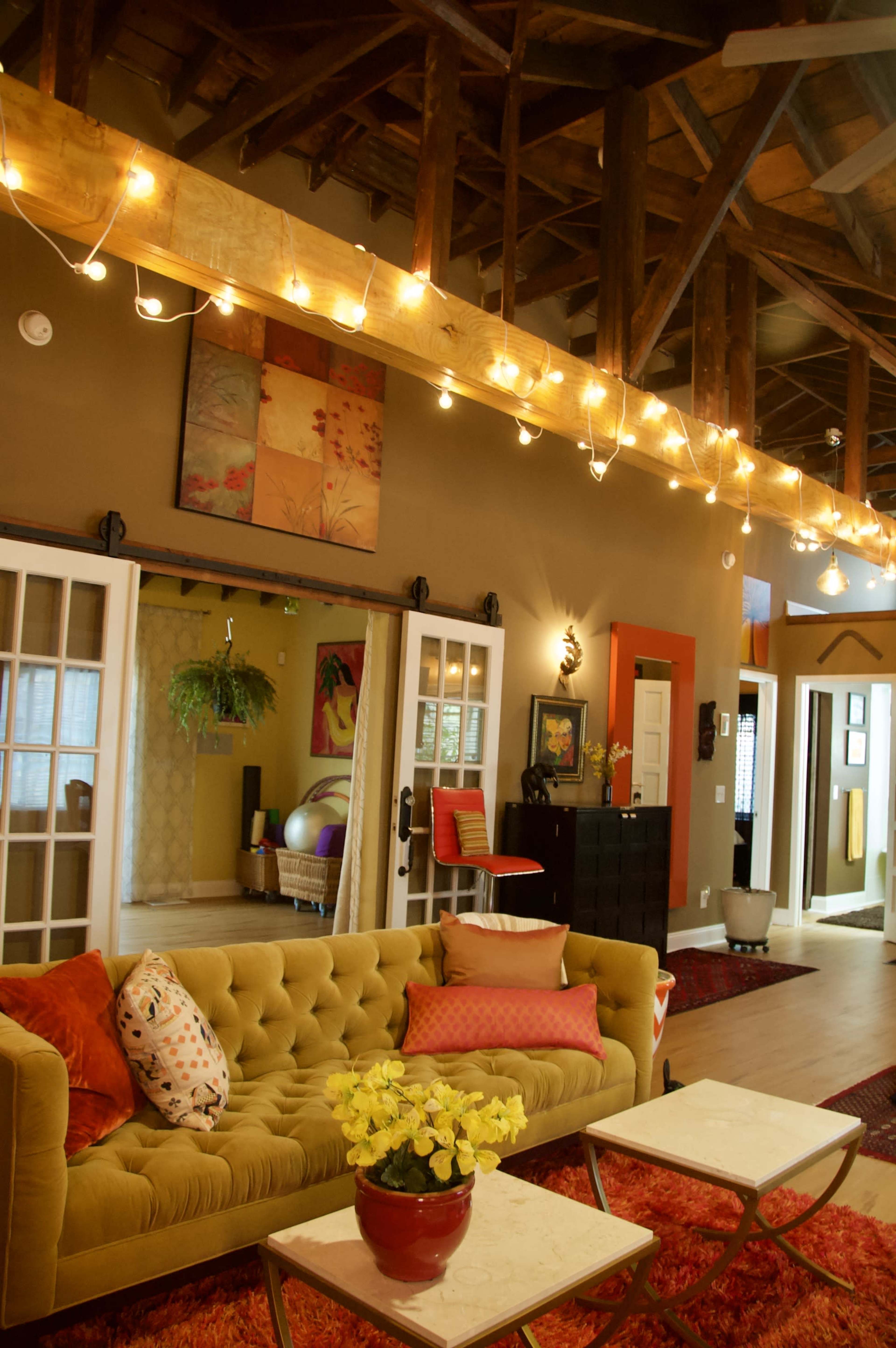 A cozy living room features a yellow velvet sofa, decorative pillows, potted flowers, and string lights illuminating the space.
