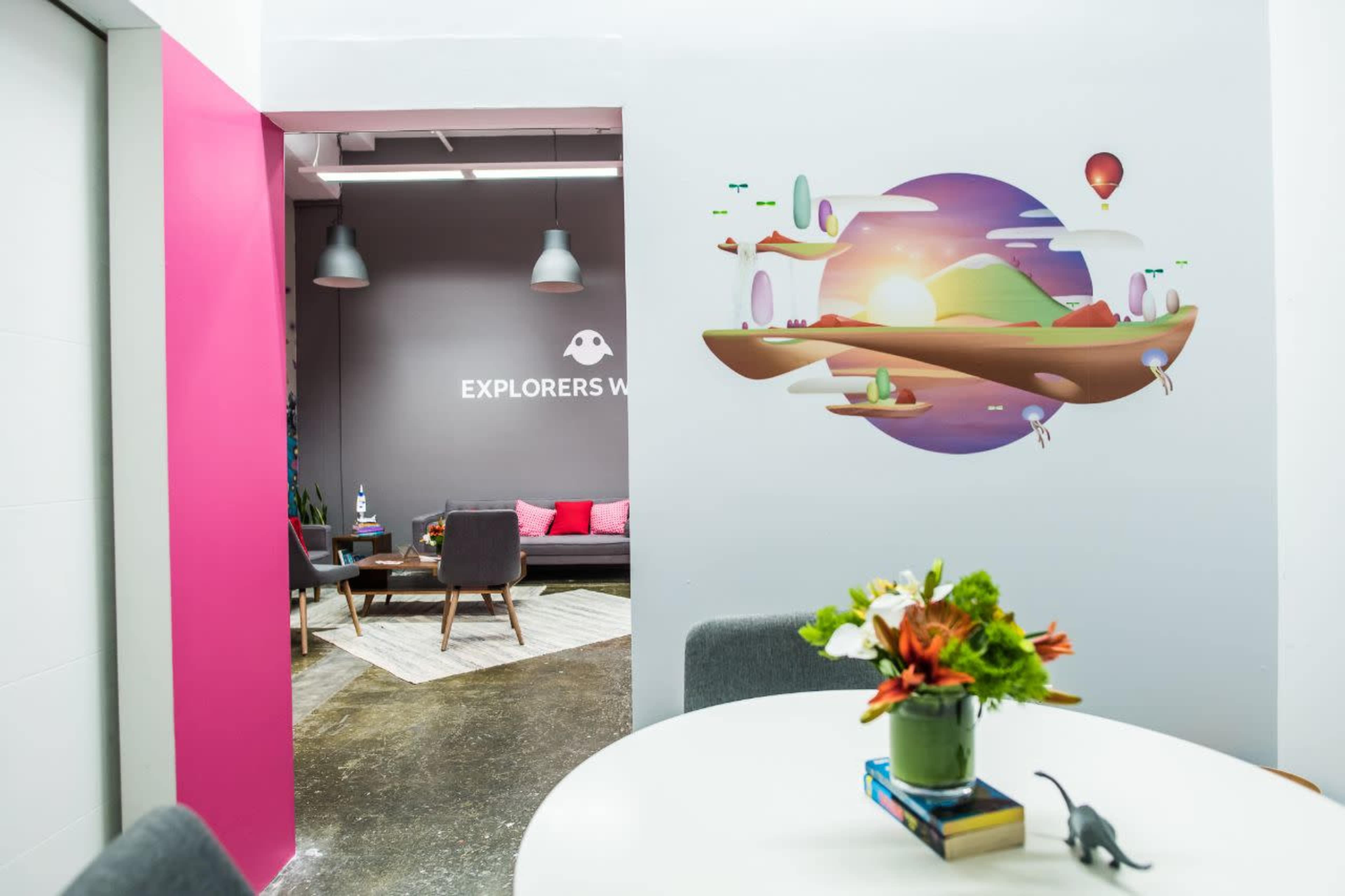 A modern office space with a brightly colored wall featuring a whimsical mural and a view into a seating area with pink accents.