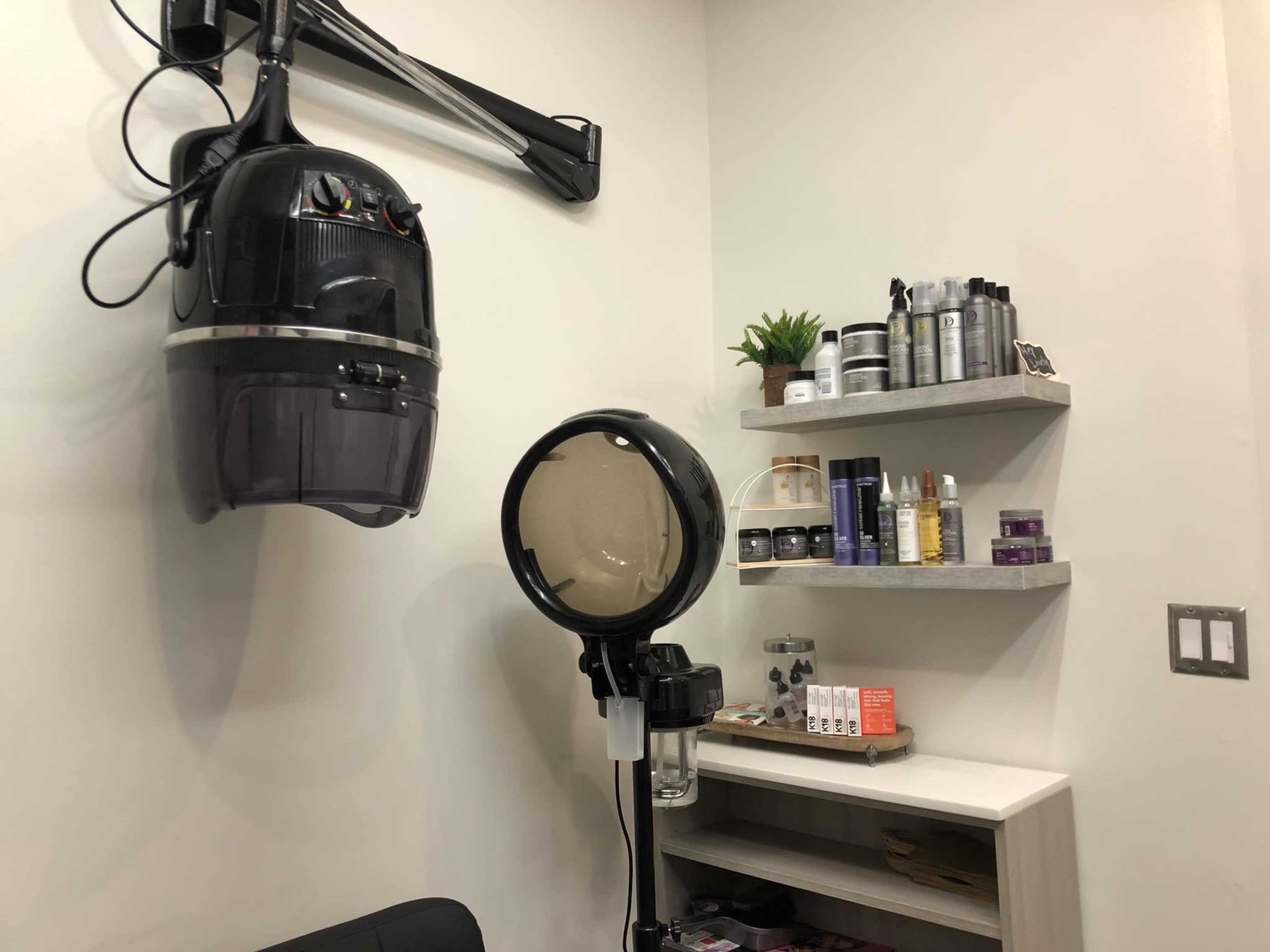 A hair salon corner featuring a hooded hair dryer mounted on the wall and a shelf filled with various hair care products.