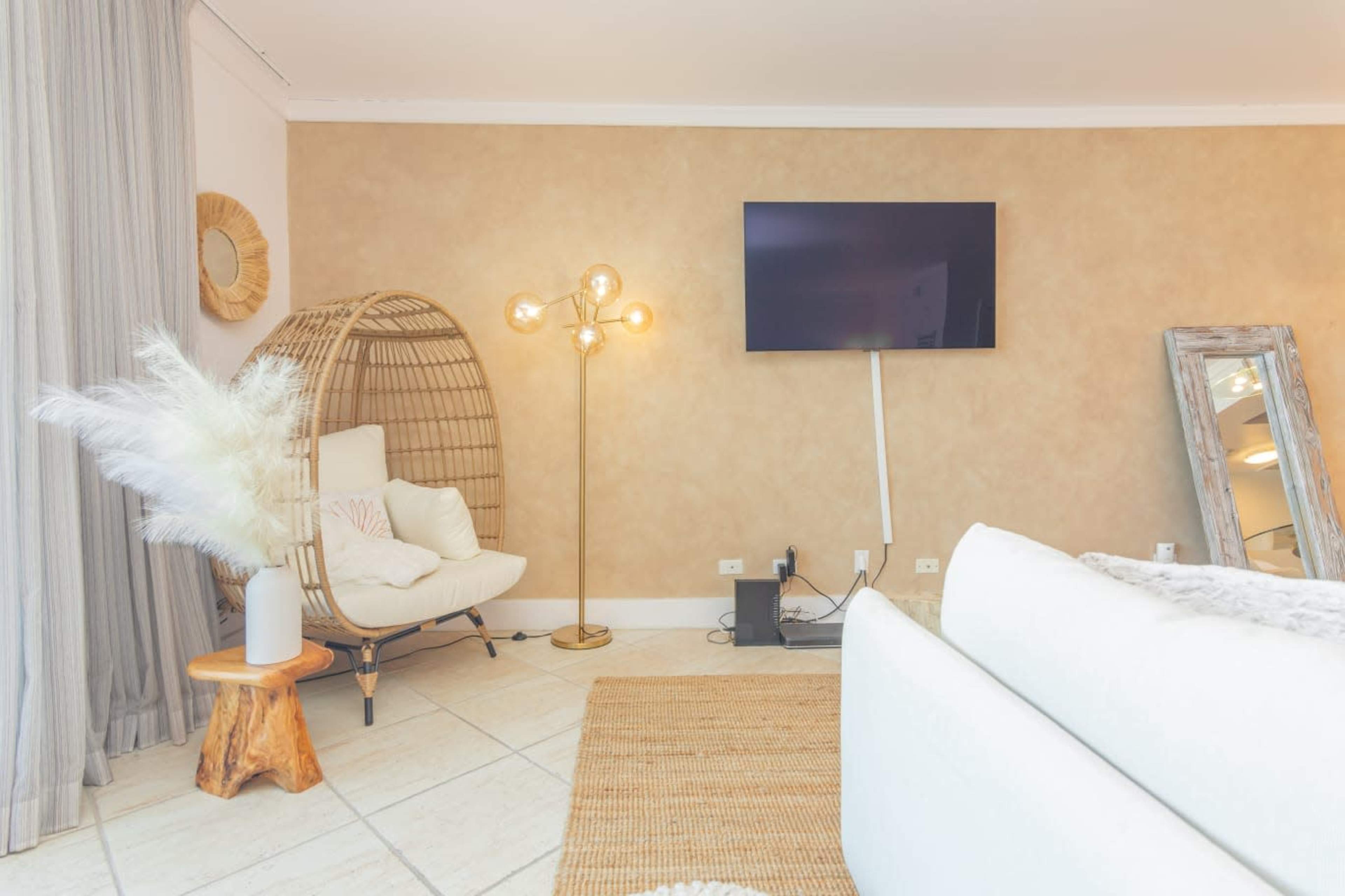 A modern living room featuring a woven chair next to a floor lamp, a wall-mounted TV, a large mirror, and decorative plants, positioned on tiled flooring.