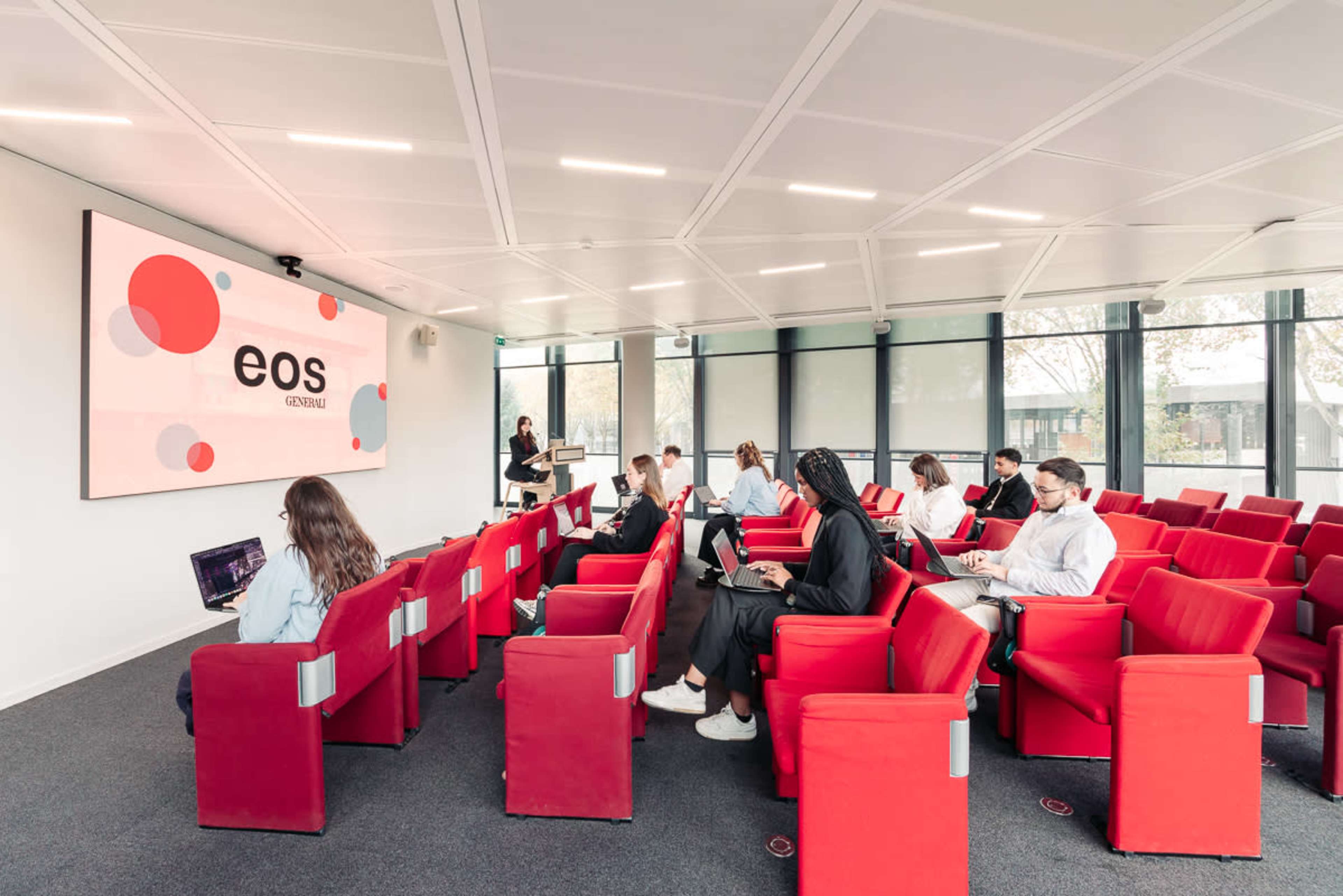 A modern conference room features red chairs arranged in rows with several individuals working on laptops as a presenter stands near a screen displaying the word "eos."
