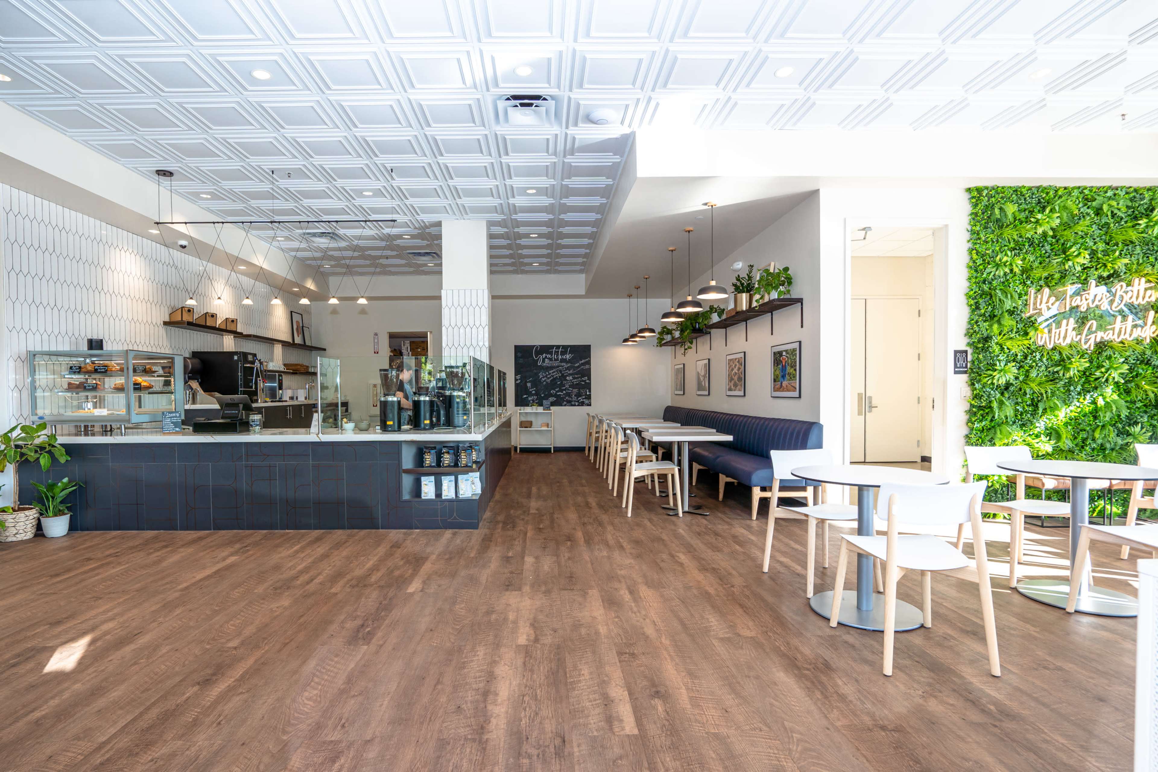 A modern café interior featuring a drinks counter, seating areas with wooden tables and chairs, and a wall with greenery.