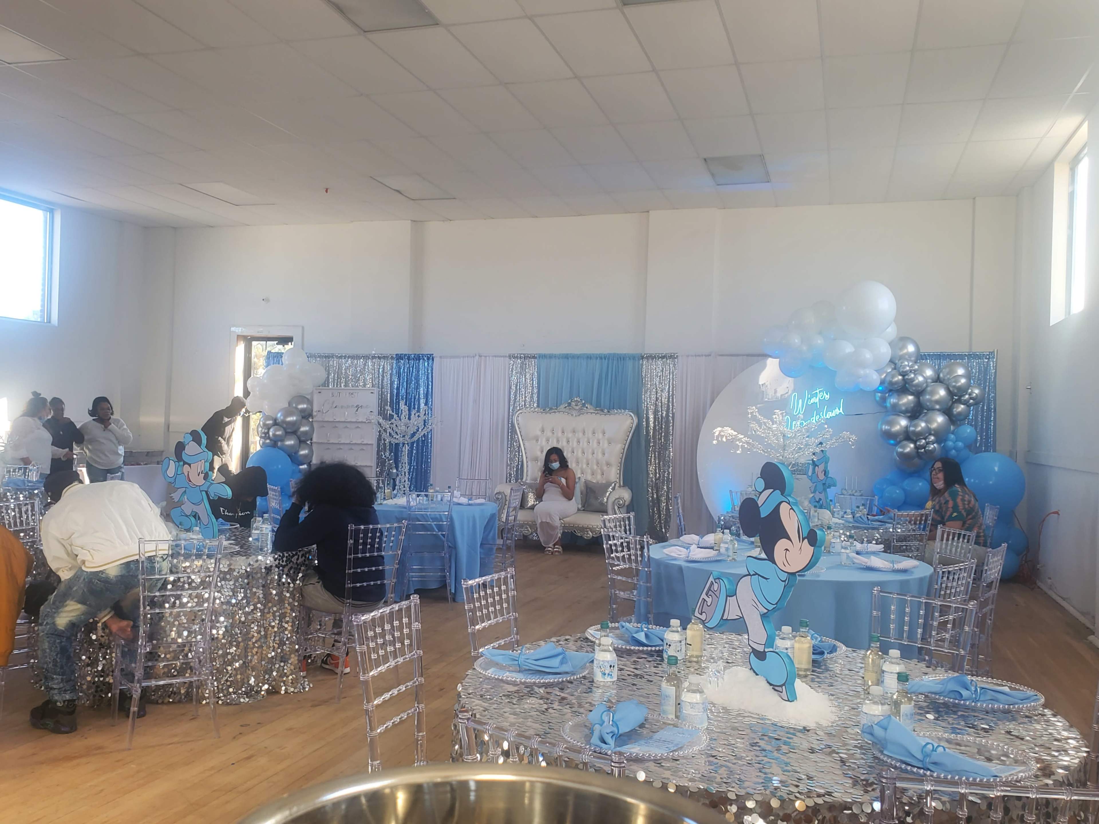 A decorated indoor venue set up for a celebration, featuring silver and blue decor, tables with sequined covers, and a large white throne chair.