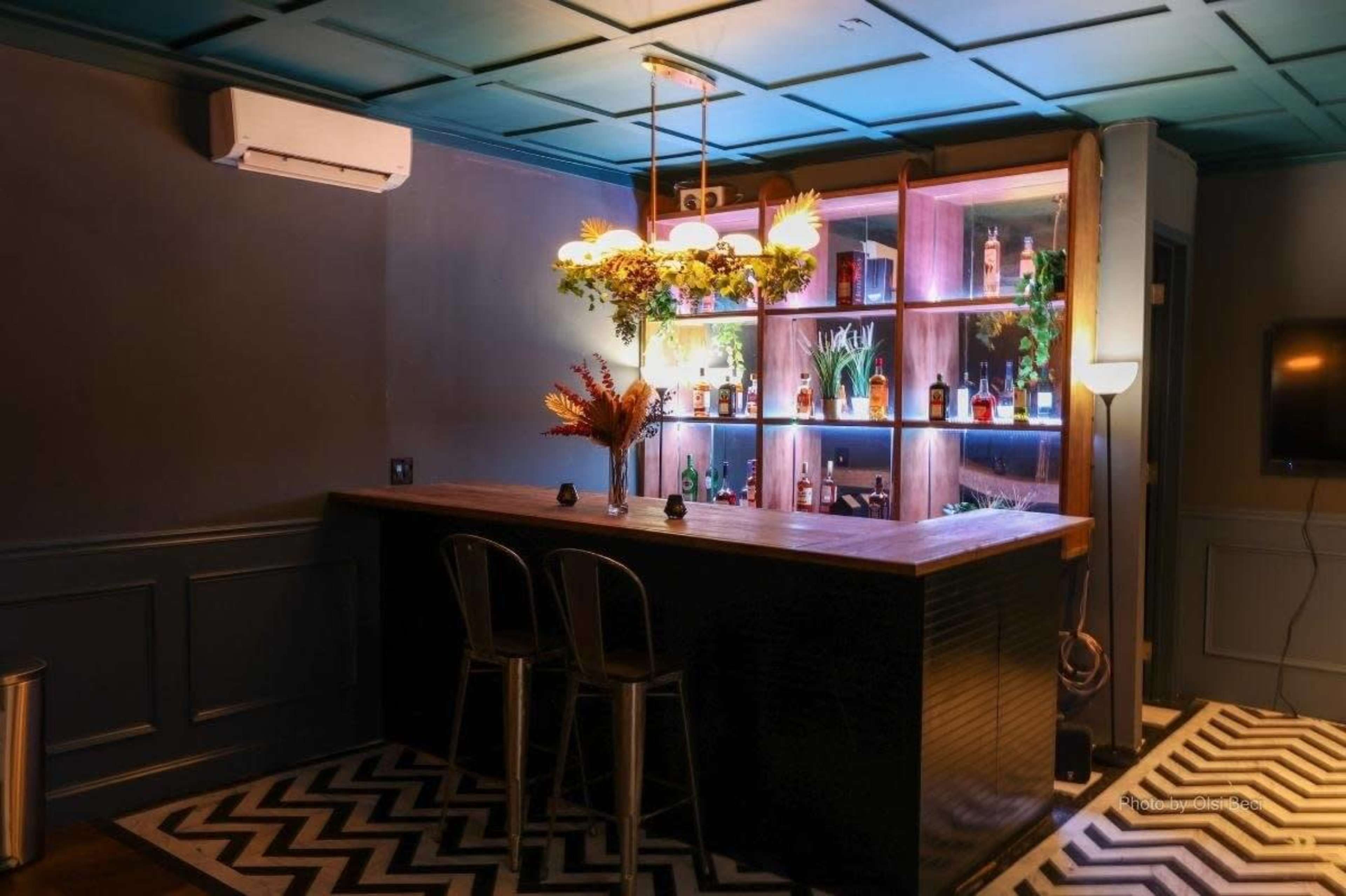 The image shows a modern bar area with a wooden counter, two metal stools, and illuminated shelves displaying various bottles.