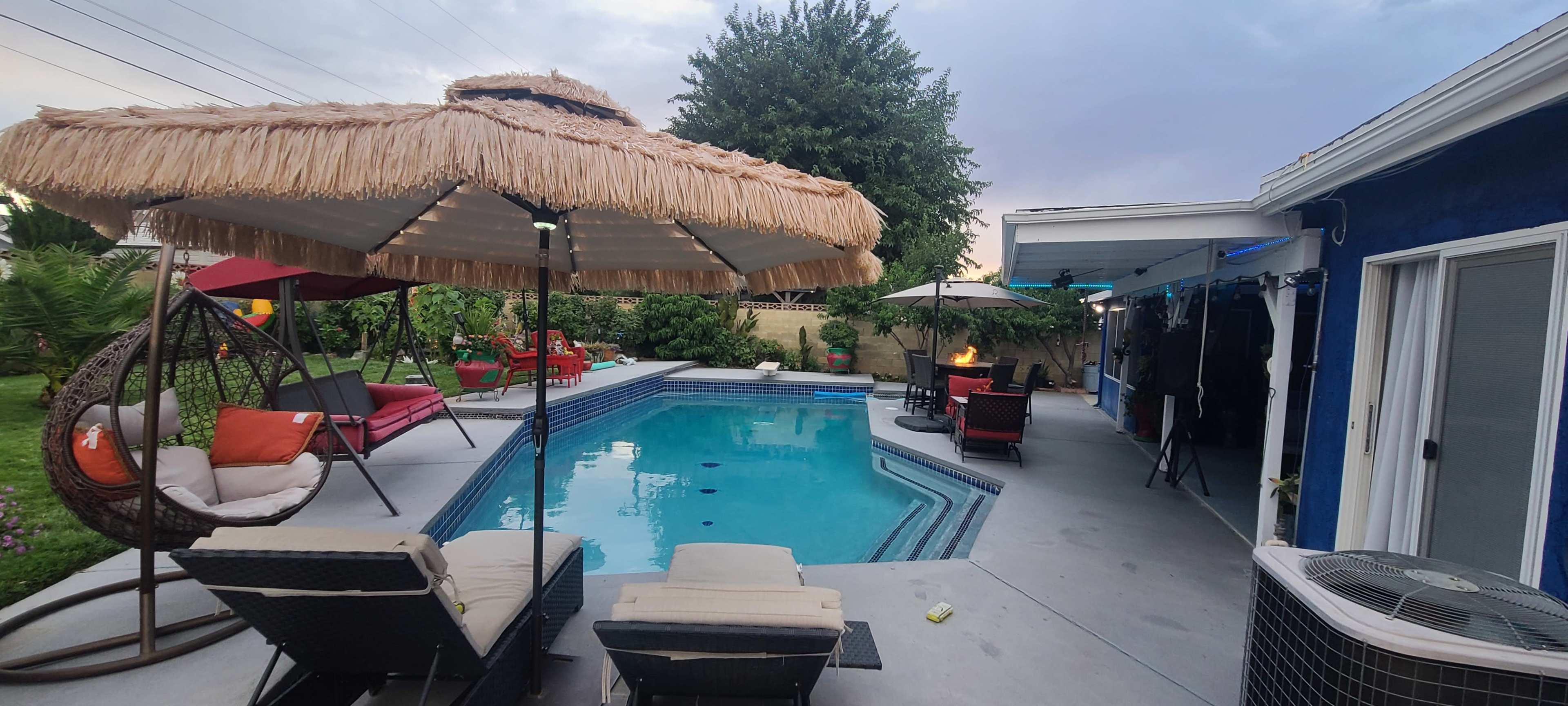 The image shows a backyard pool area featuring lounge chairs, umbrellas, and various seating arrangements surrounded by greenery and a house.