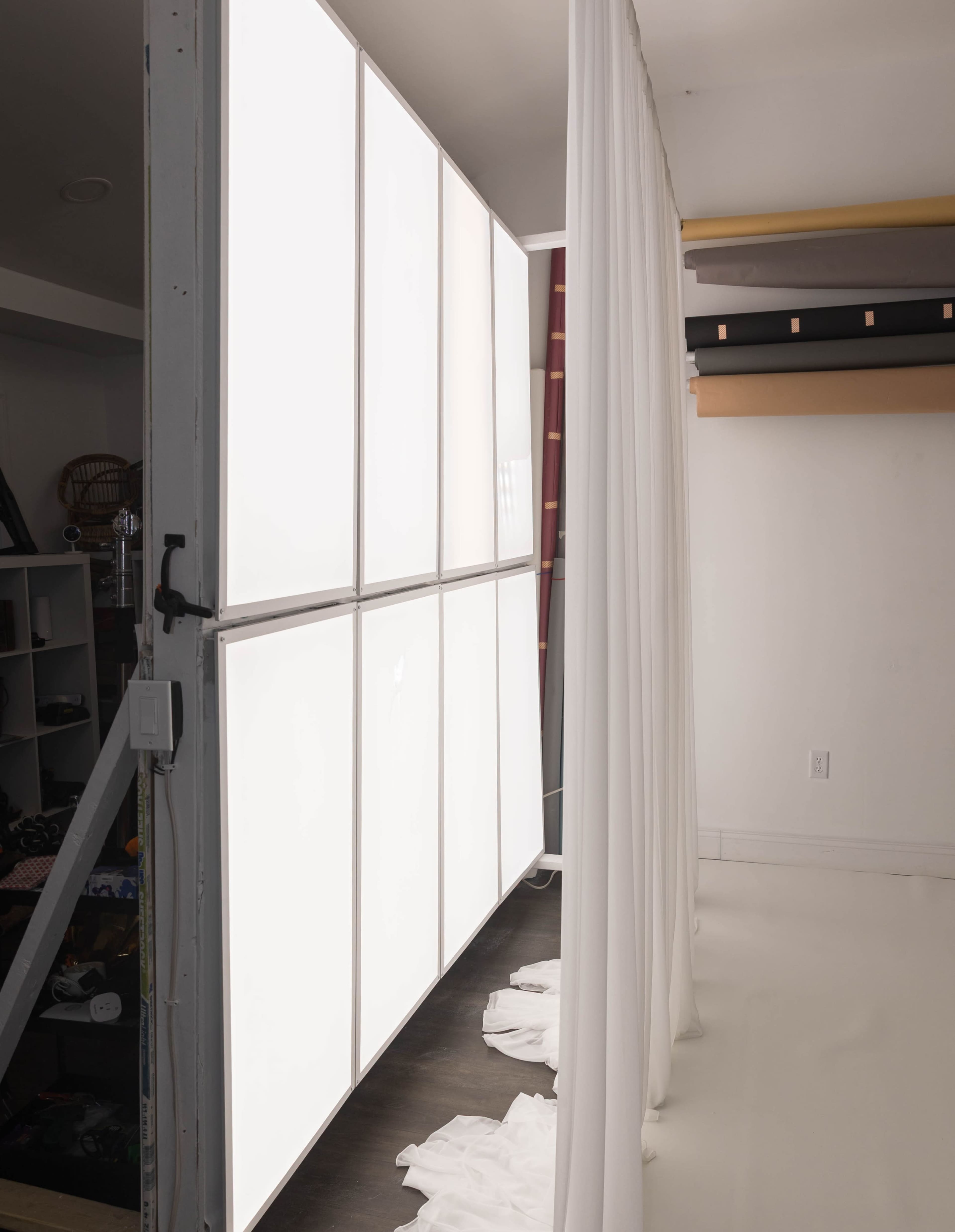 The image shows a bright, panelled light source against a wall, with a white curtain partially draping in front of it on a dark floor.