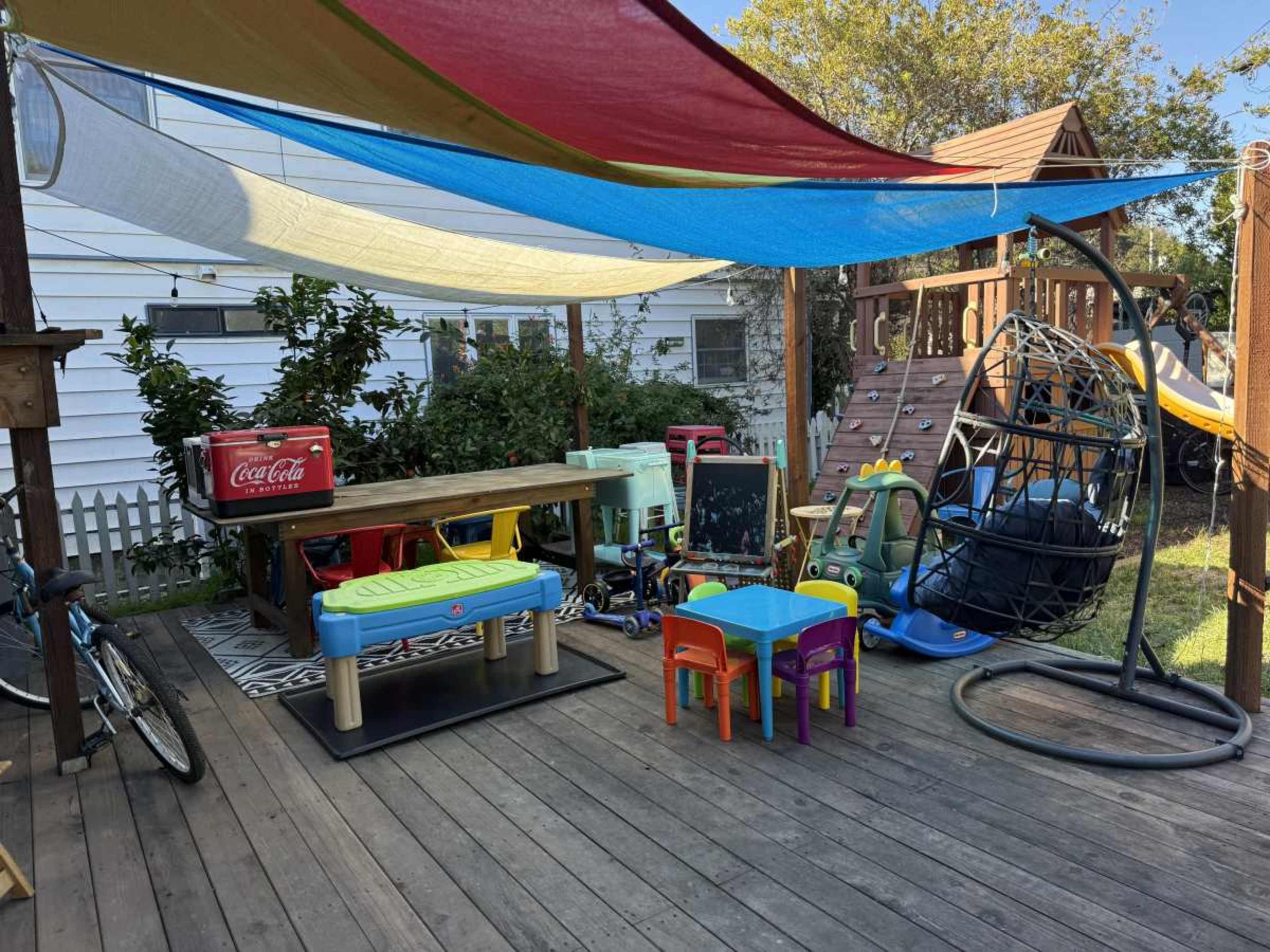 A backyard play area features colorful shade sails above a wooden deck with children’s toys, a small table and chairs, and a bicycle.