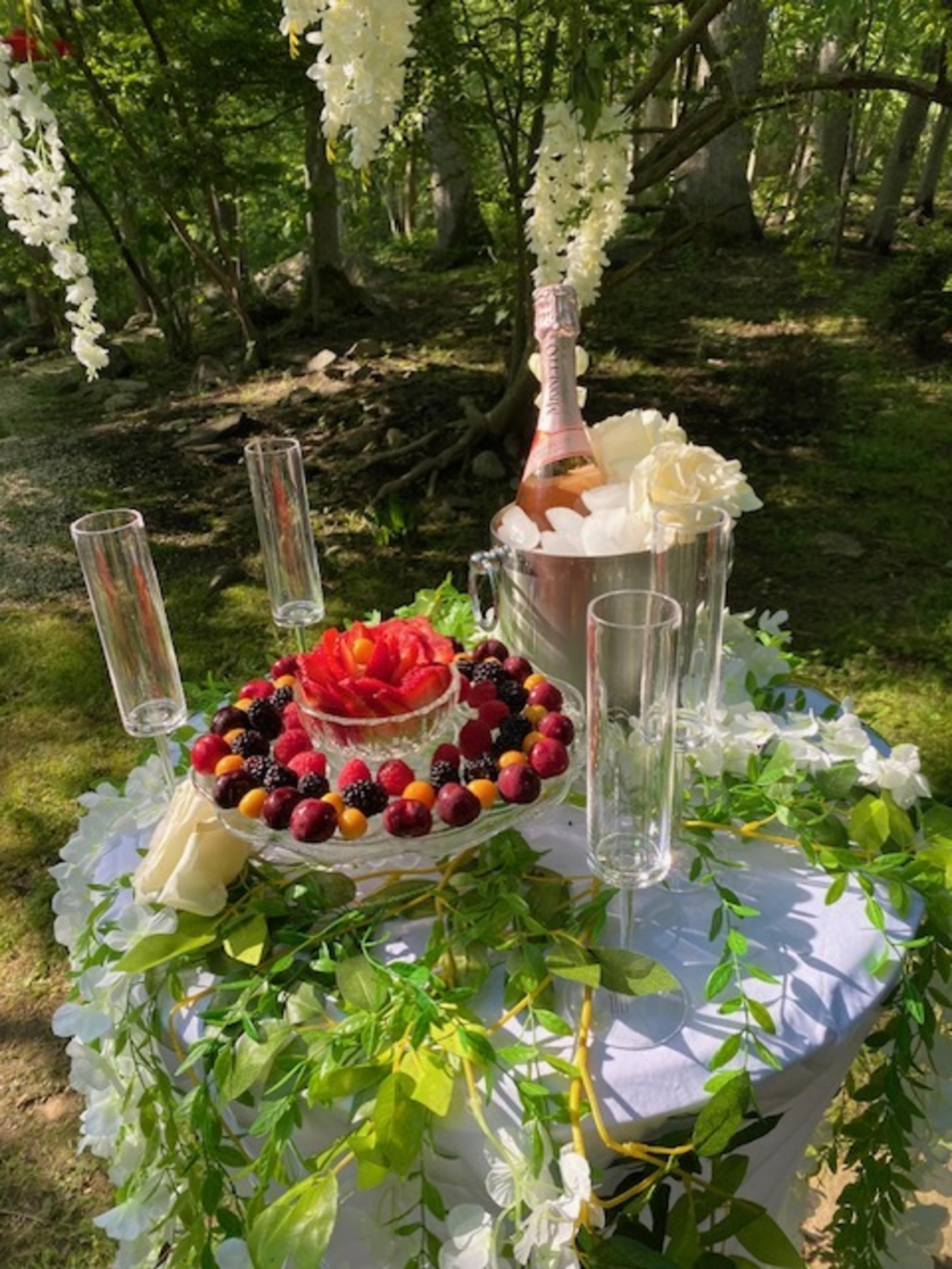 A decorated table features a fruit arrangement surrounded by champagne glasses and a bottle of sparkling wine in a woodland setting.
