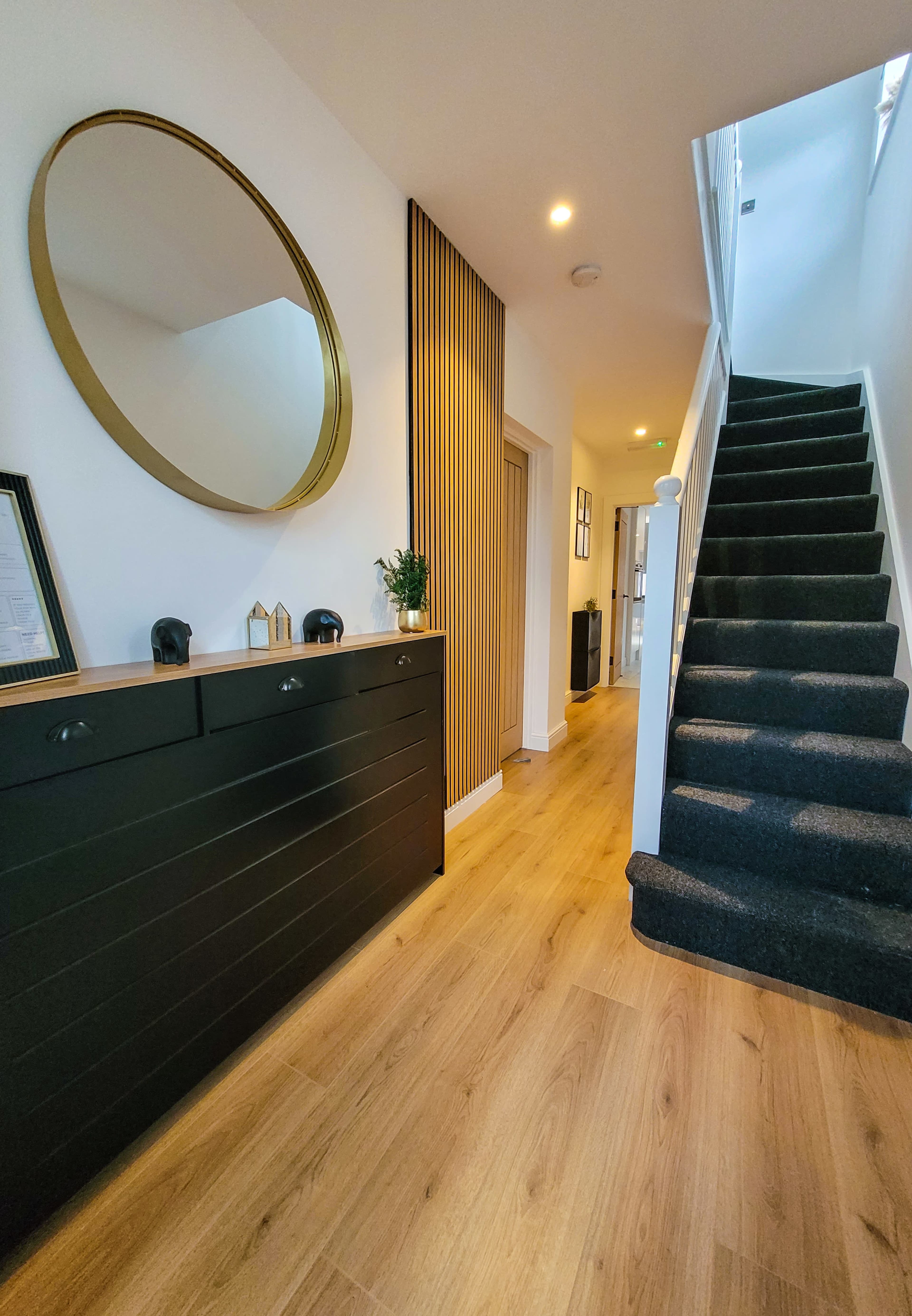 A modern hallway featuring a round mirror above a black console table, wooden flooring, and a staircase leading to the upper level.