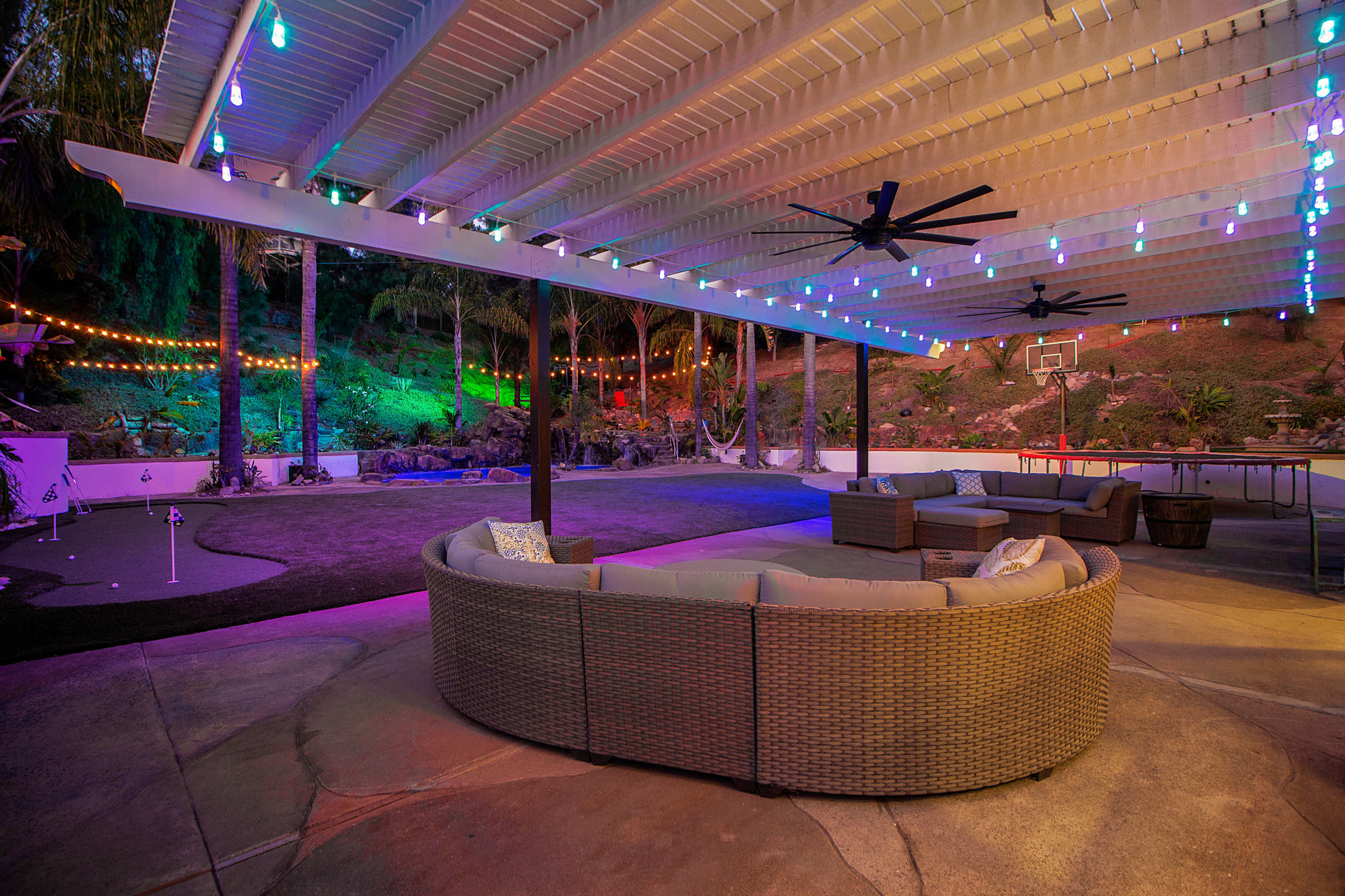 A spacious outdoor lounge area with a circular sectional sofa under a covered patio, featuring colorful string lights and a basketball hoop in the background.
