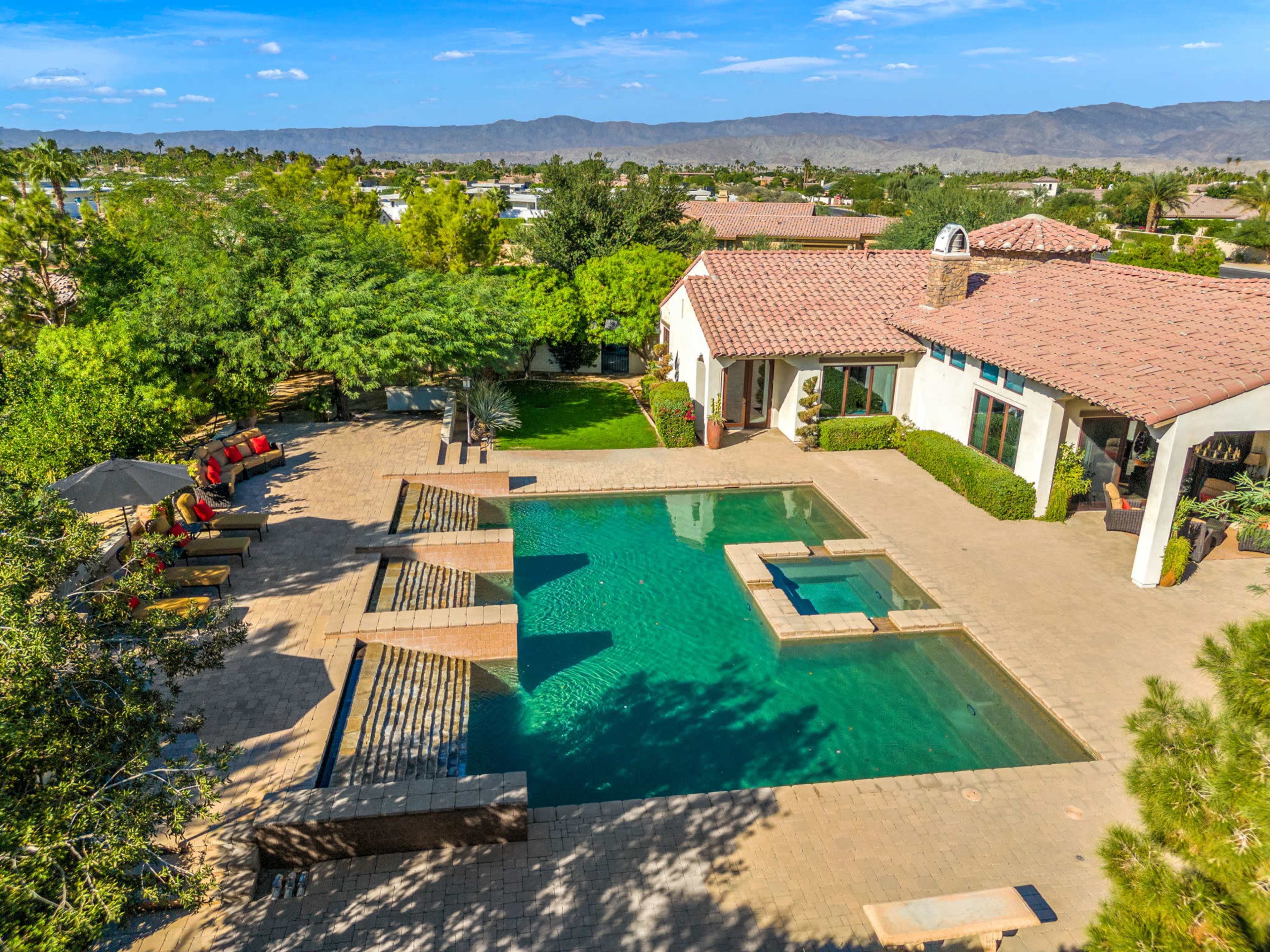 A spacious backyard with a swimming pool, hot tub, and lounge chairs, surrounded by landscaped greenery and a house with a tiled roof.