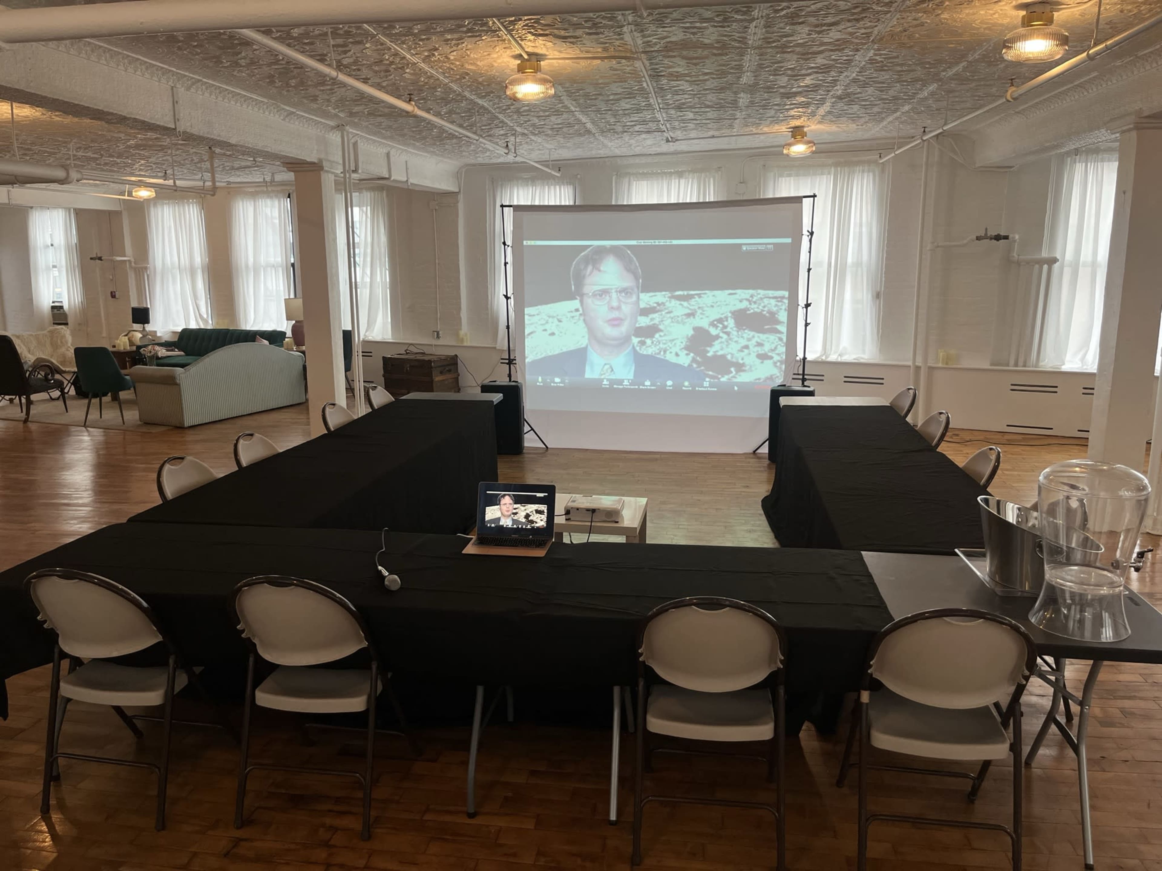 A conference room set up with eight black tables arranged in a U-shape, a projector screen displaying a video, and chairs placed around the tables.
