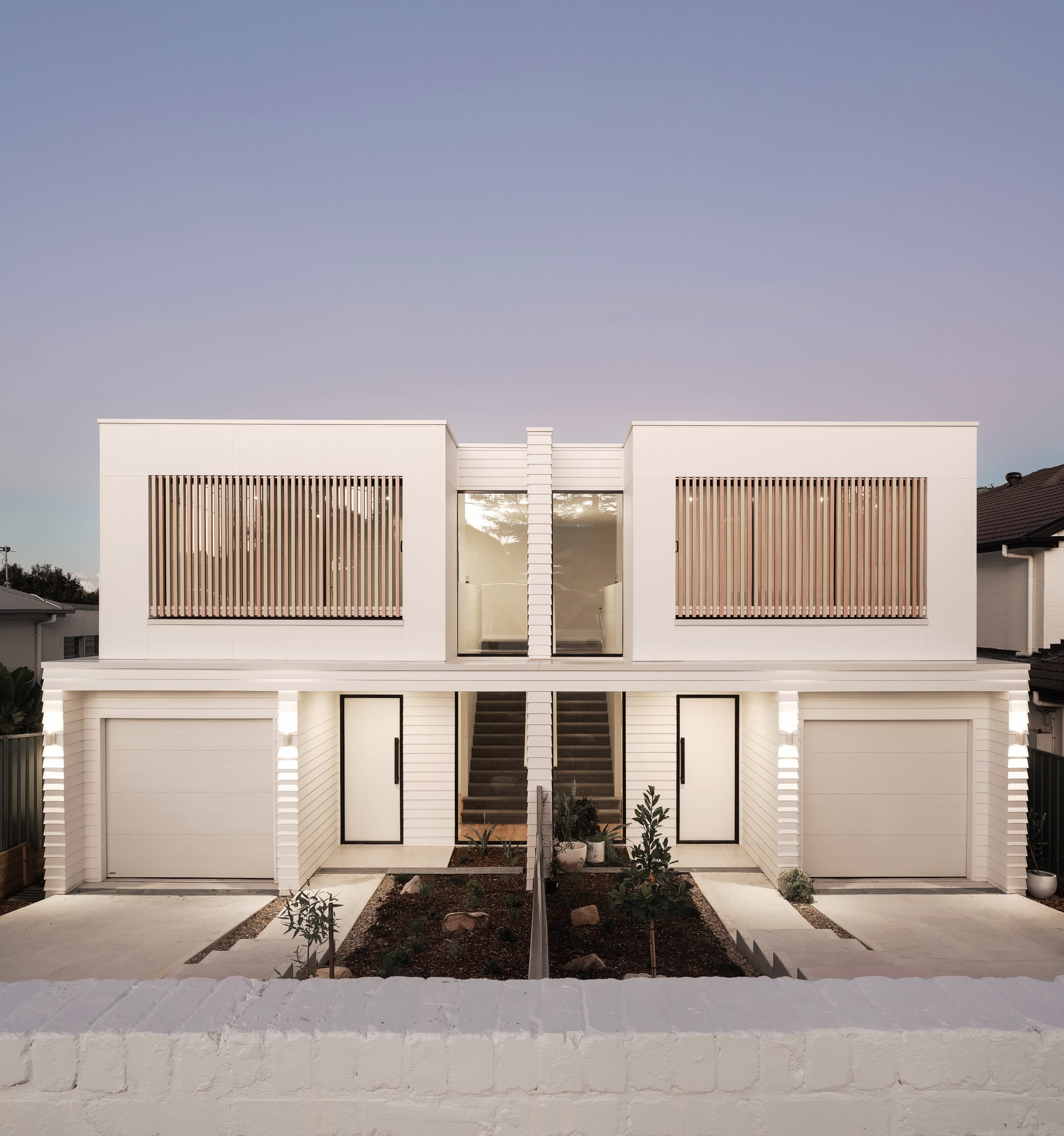 The image shows a modern two-story house with a symmetrical façade, featuring horizontal wooden slats over large windows and a landscaped pathway leading to the entrance.