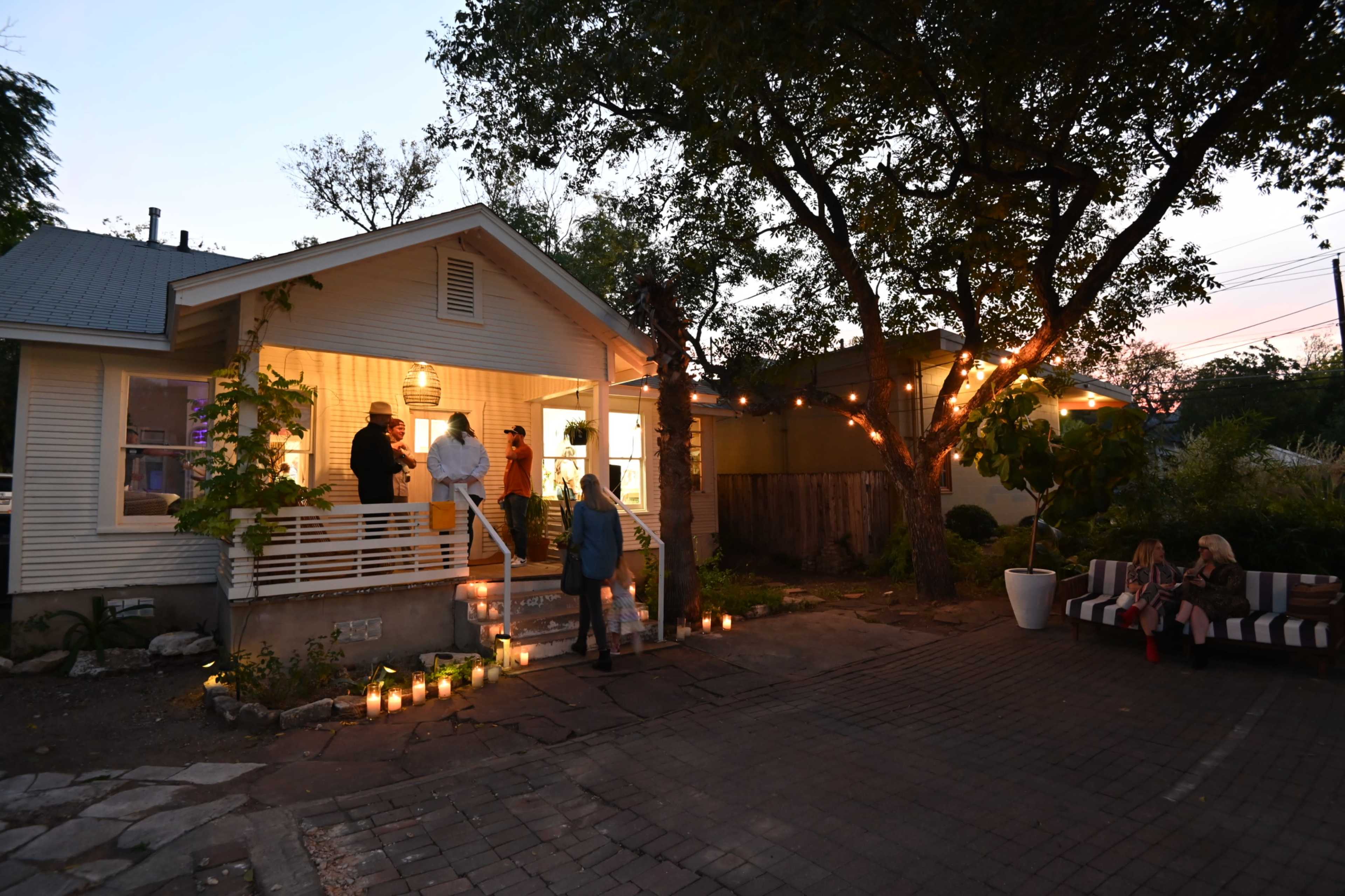 A group of people gathers on the porch of a well-lit house with candles lining the pathway as twilight approaches.