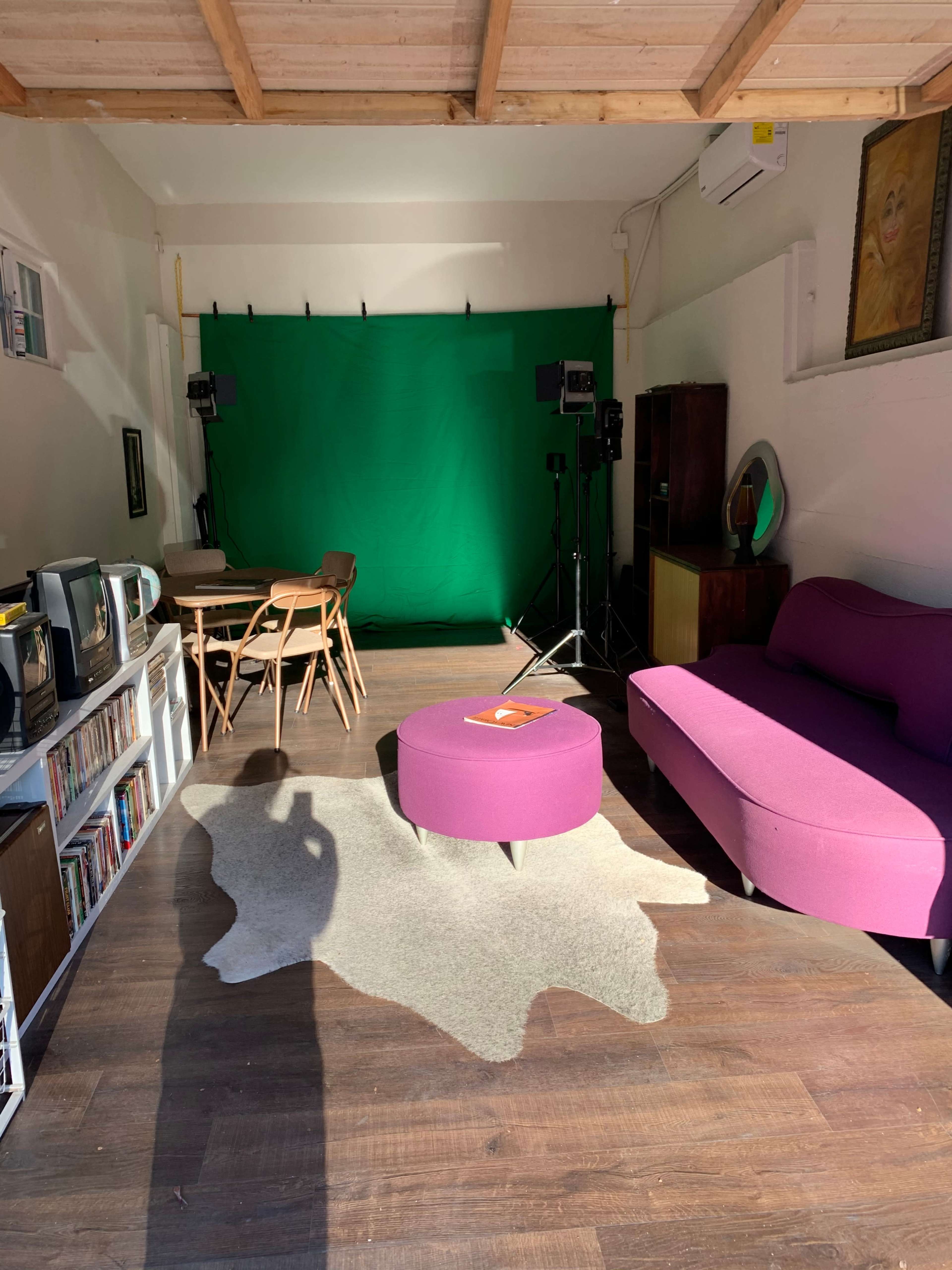 A modern room features a green backdrop with studio lights, a pink sofa, a circular purple coffee table, and a wooden dining set.