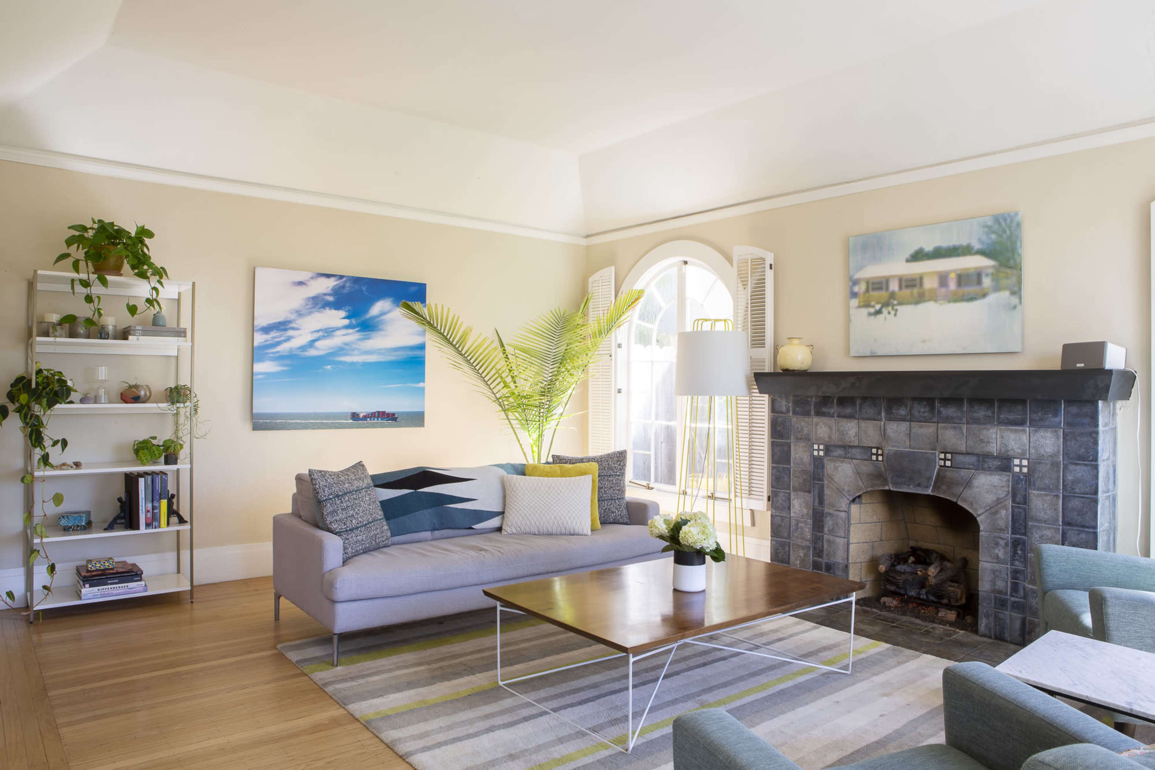A modern living room features a gray sofa, a wooden coffee table, a stone fireplace, and large windows with a view, complemented by colorful wall art and indoor plants.