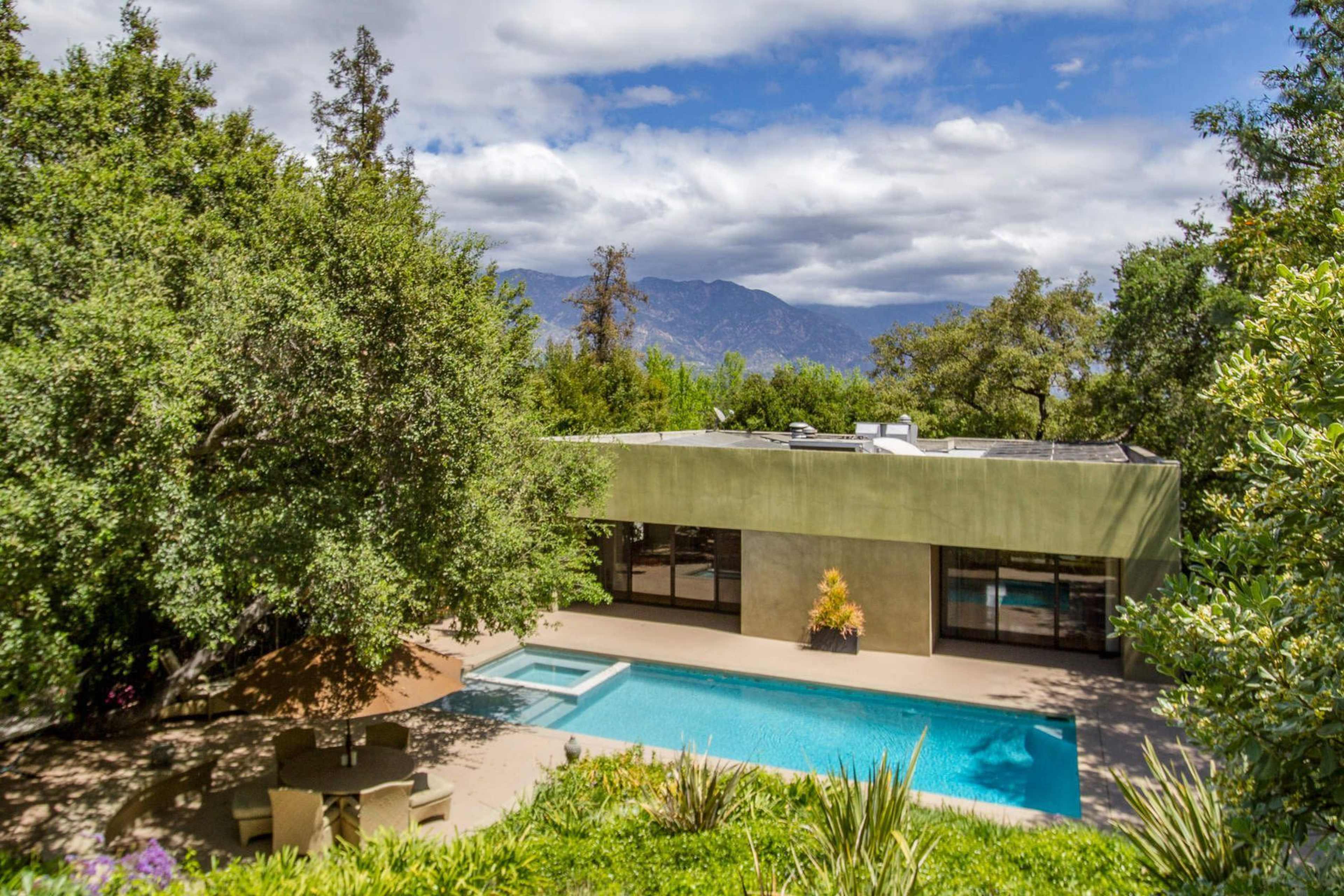 A modern house with large windows overlooks a swimming pool surrounded by greenery and mountains in the background.