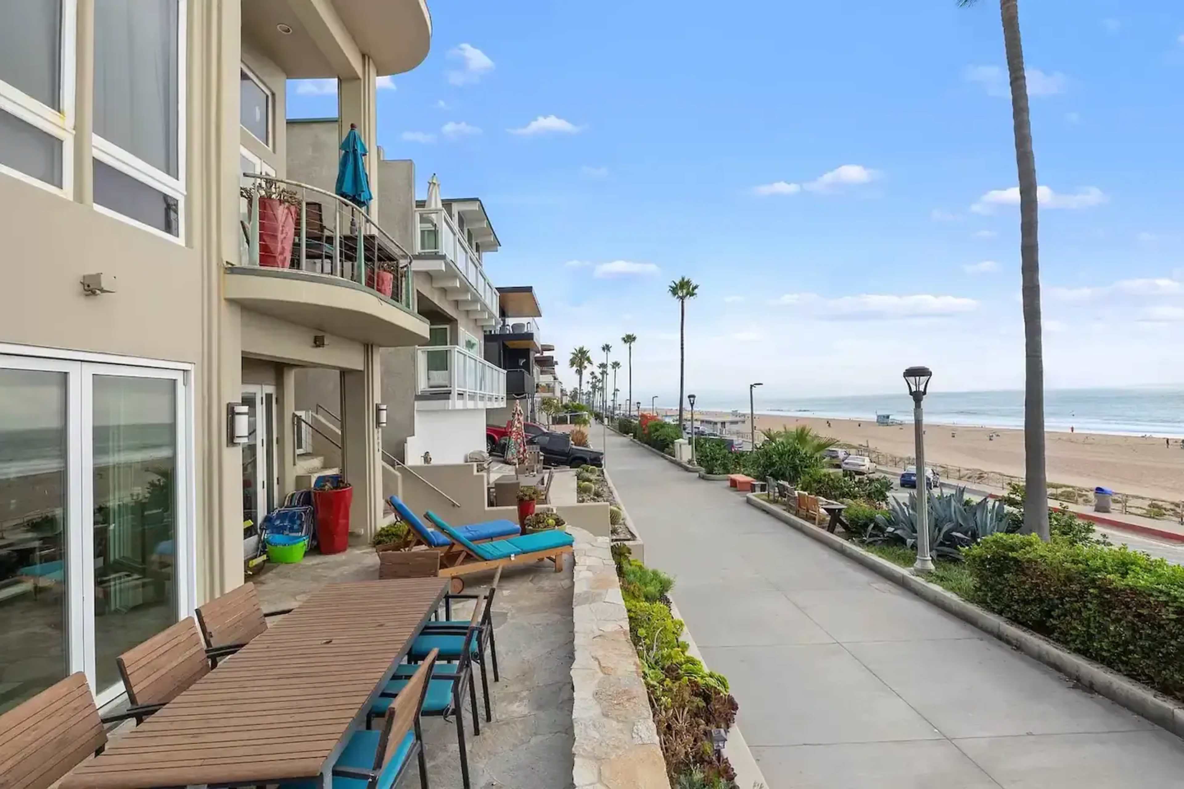 A beachfront walkway lined with palm trees and residential buildings, with outdoor seating and a view of the ocean in the distance.