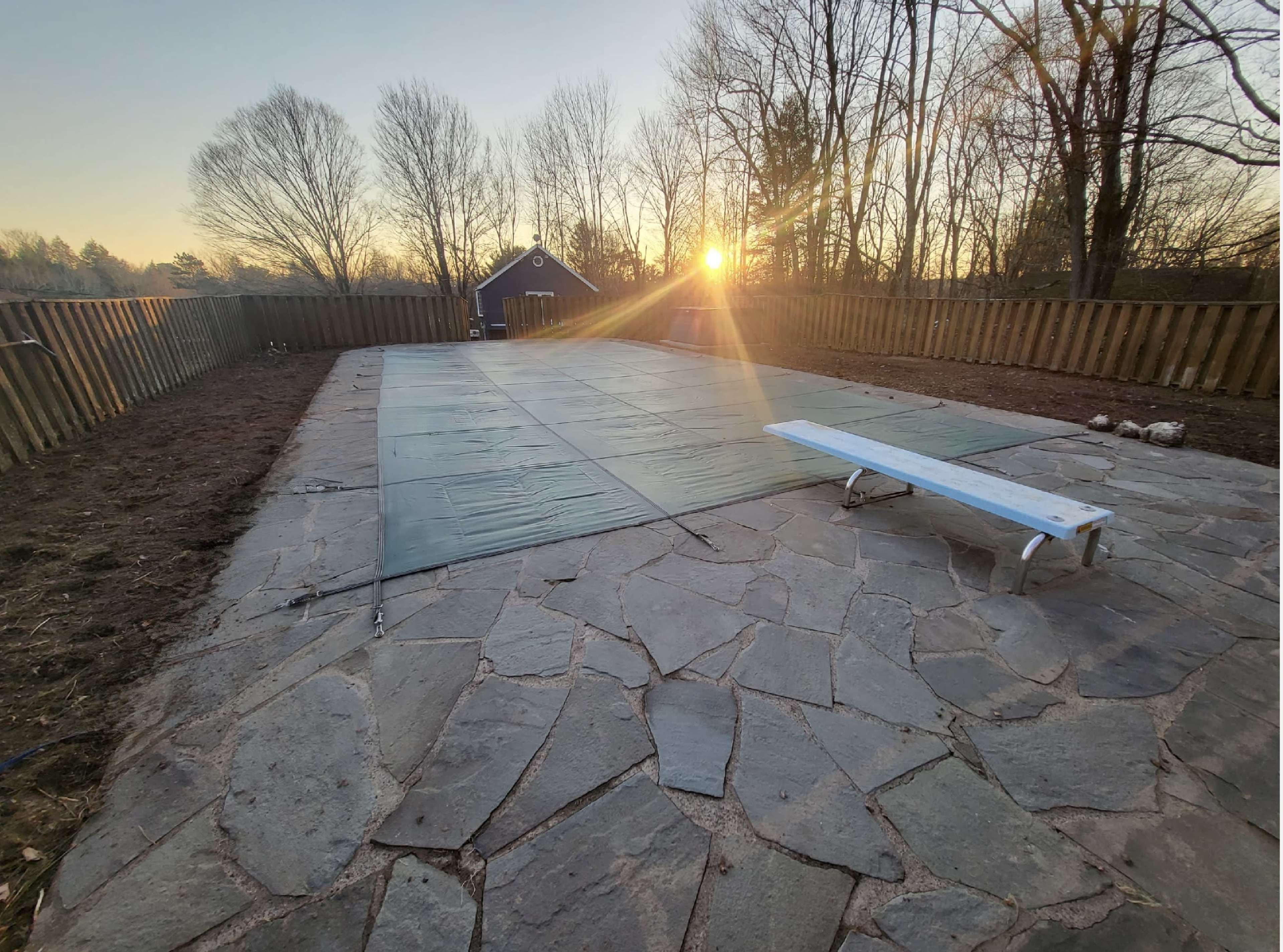 A pool area covered with a green safety cover, surrounded by a stone patio and a wooden fence, with a setting sun in the background.
