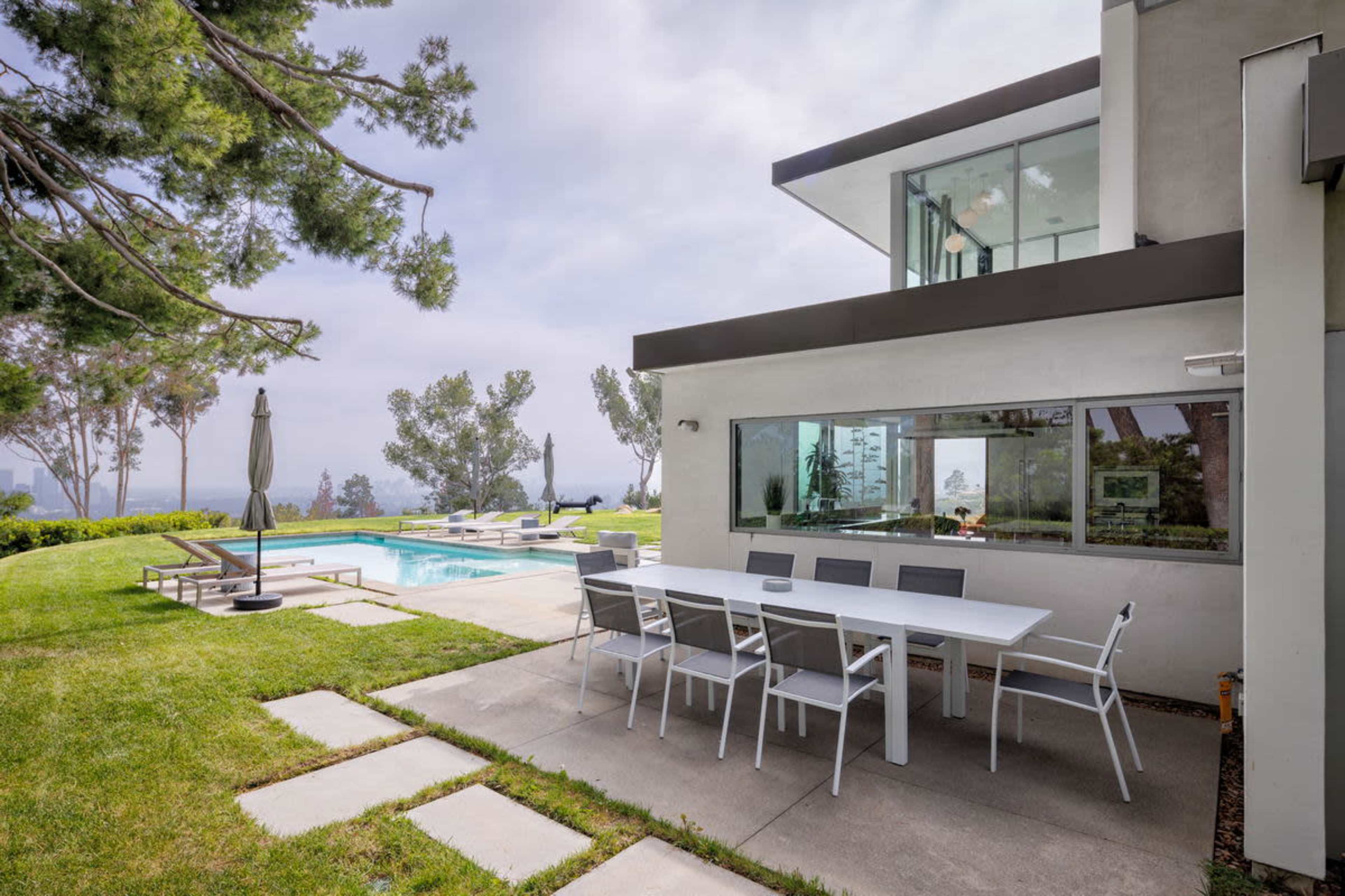 A modern home features a patio with a dining table adjacent to a swimming pool surrounded by greenery.