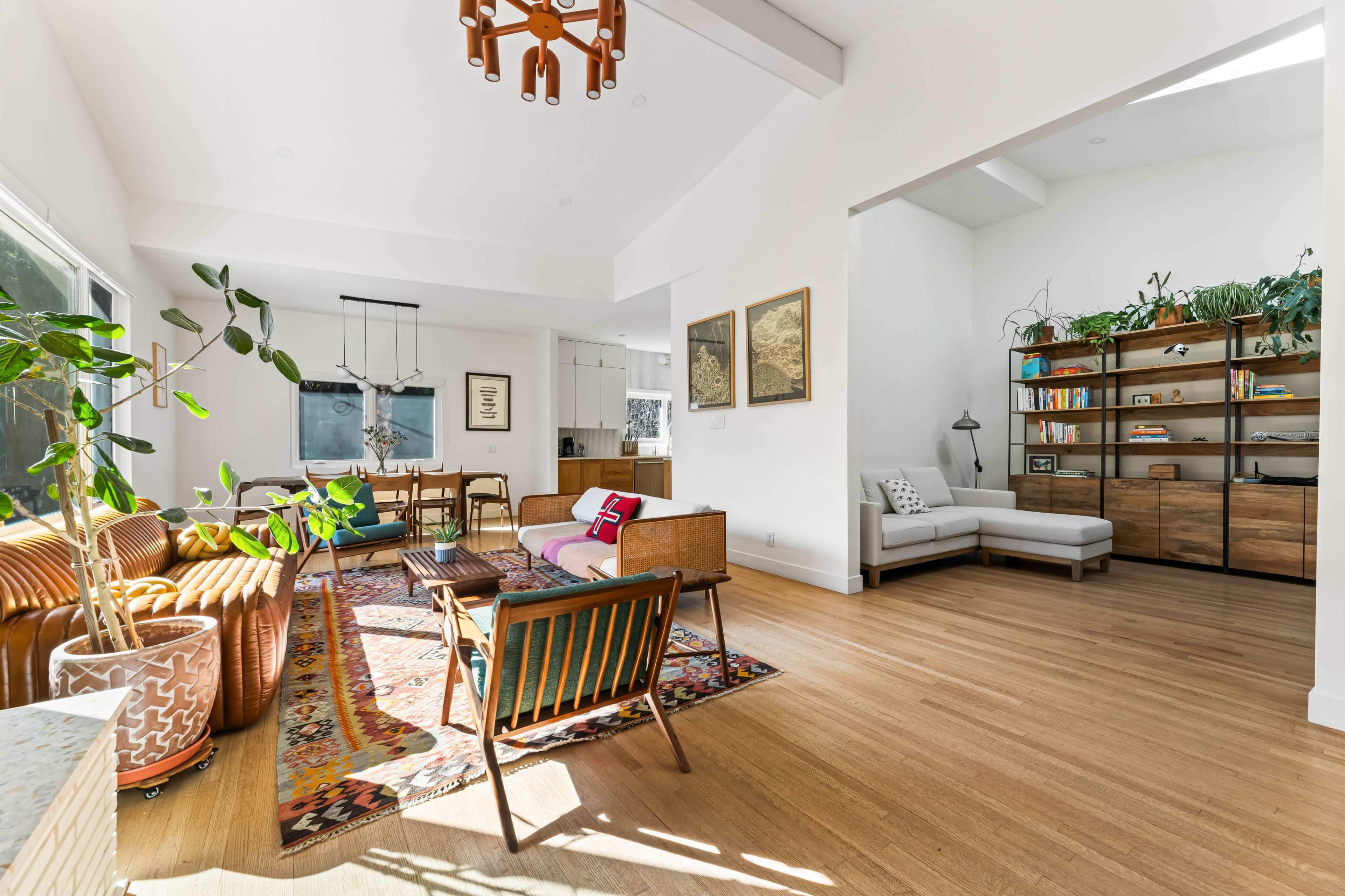 The image shows a bright, modern living space with a mix of seating areas, decorative plants, and stylish furniture arranged around a colorful area rug.