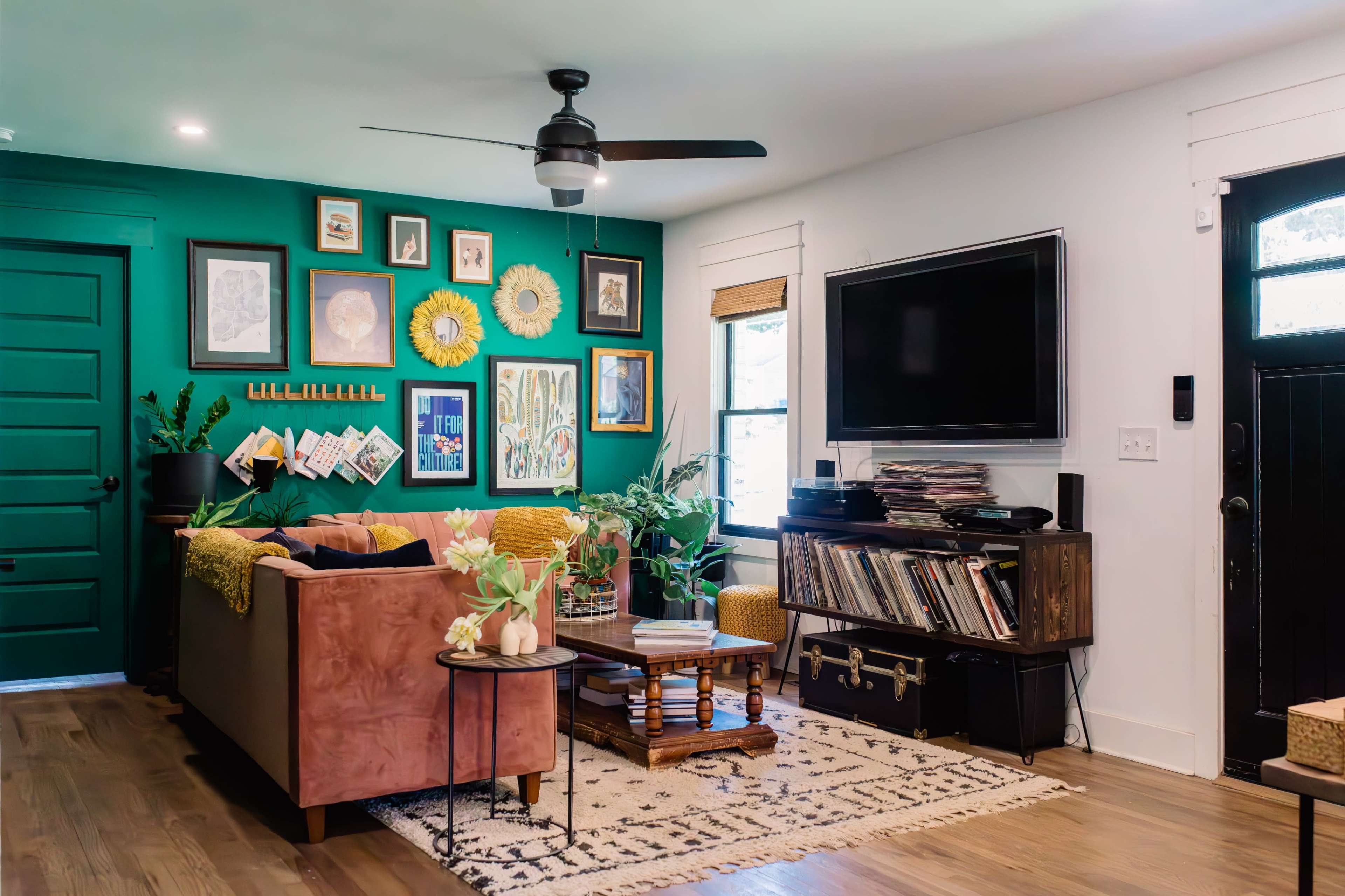 The living room features a green accent wall adorned with a gallery of framed art, a brown sofa with yellow pillows, and a media console displaying vinyl records.