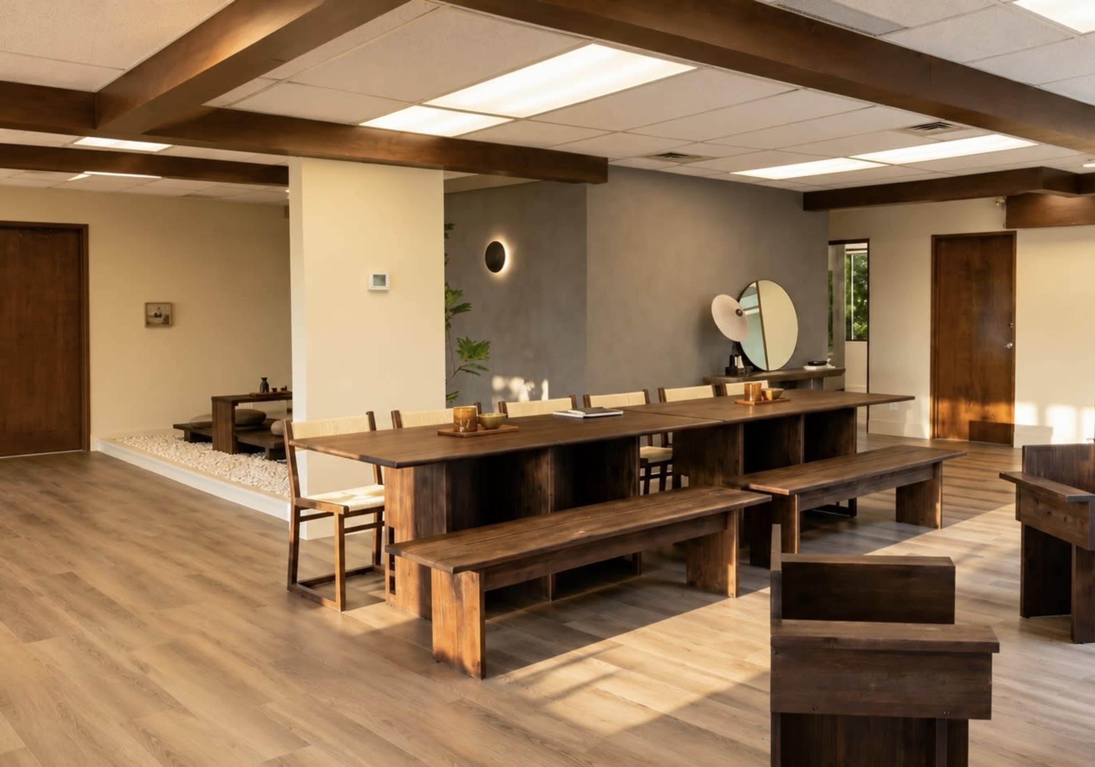 The image shows a spacious, modern room with wooden furniture, including a long table and benches, surrounded by minimalist décor and natural light.