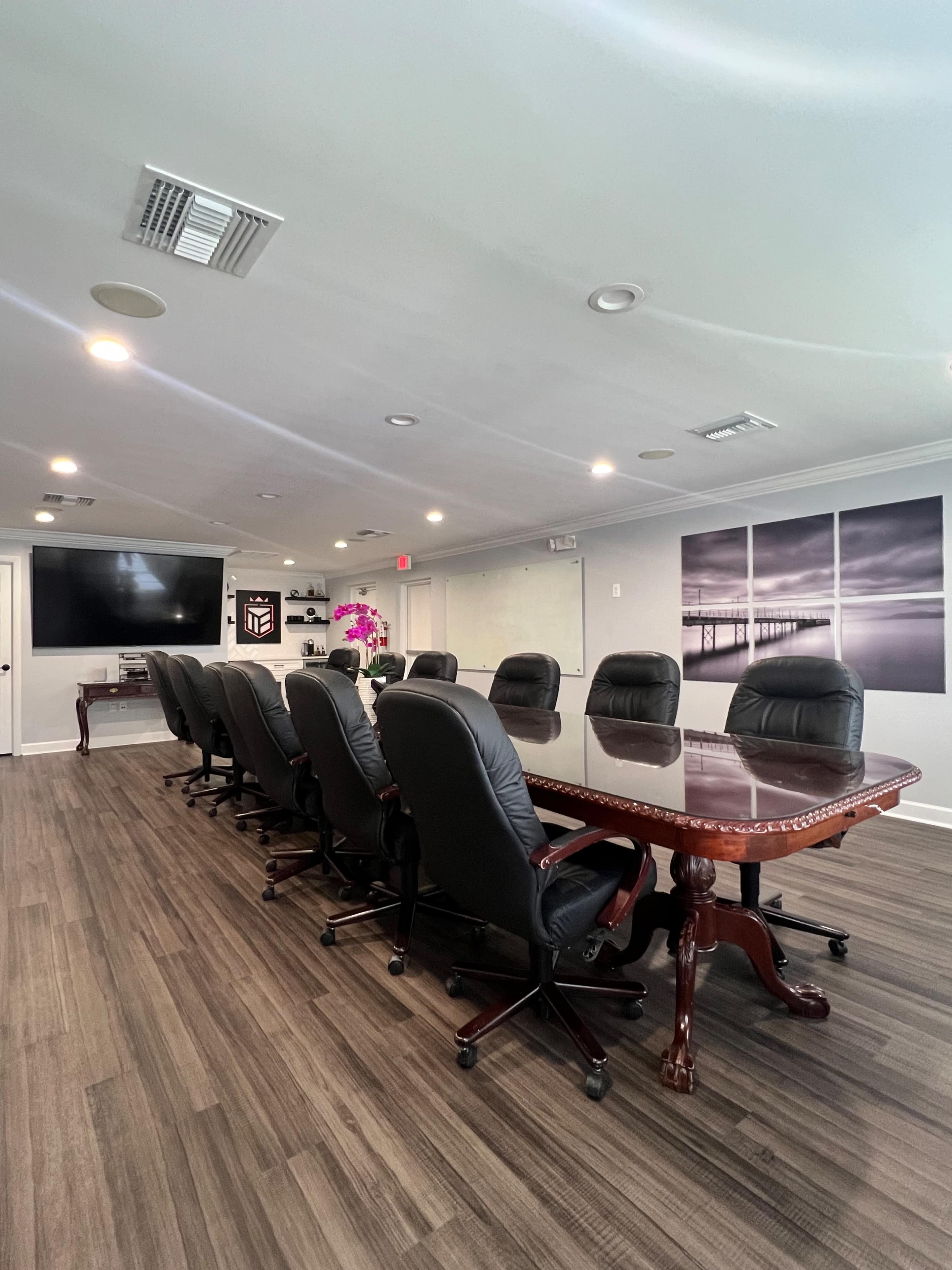 A modern conference room with a long wooden table surrounded by black rolling chairs and equipped with a large screen at one end.