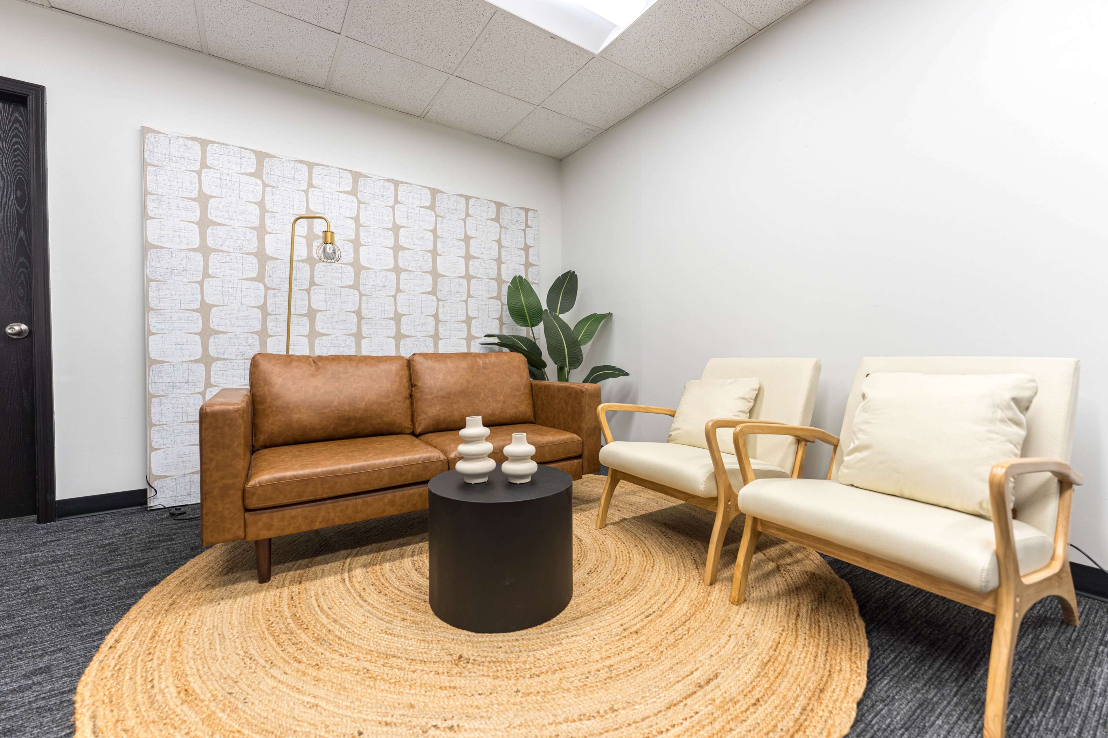 The image shows a sitting area with a brown leather sofa, two light-colored chairs, a round black table, and a circular jute rug on a gray carpet, alongside a plant and a patterned wall decoration.