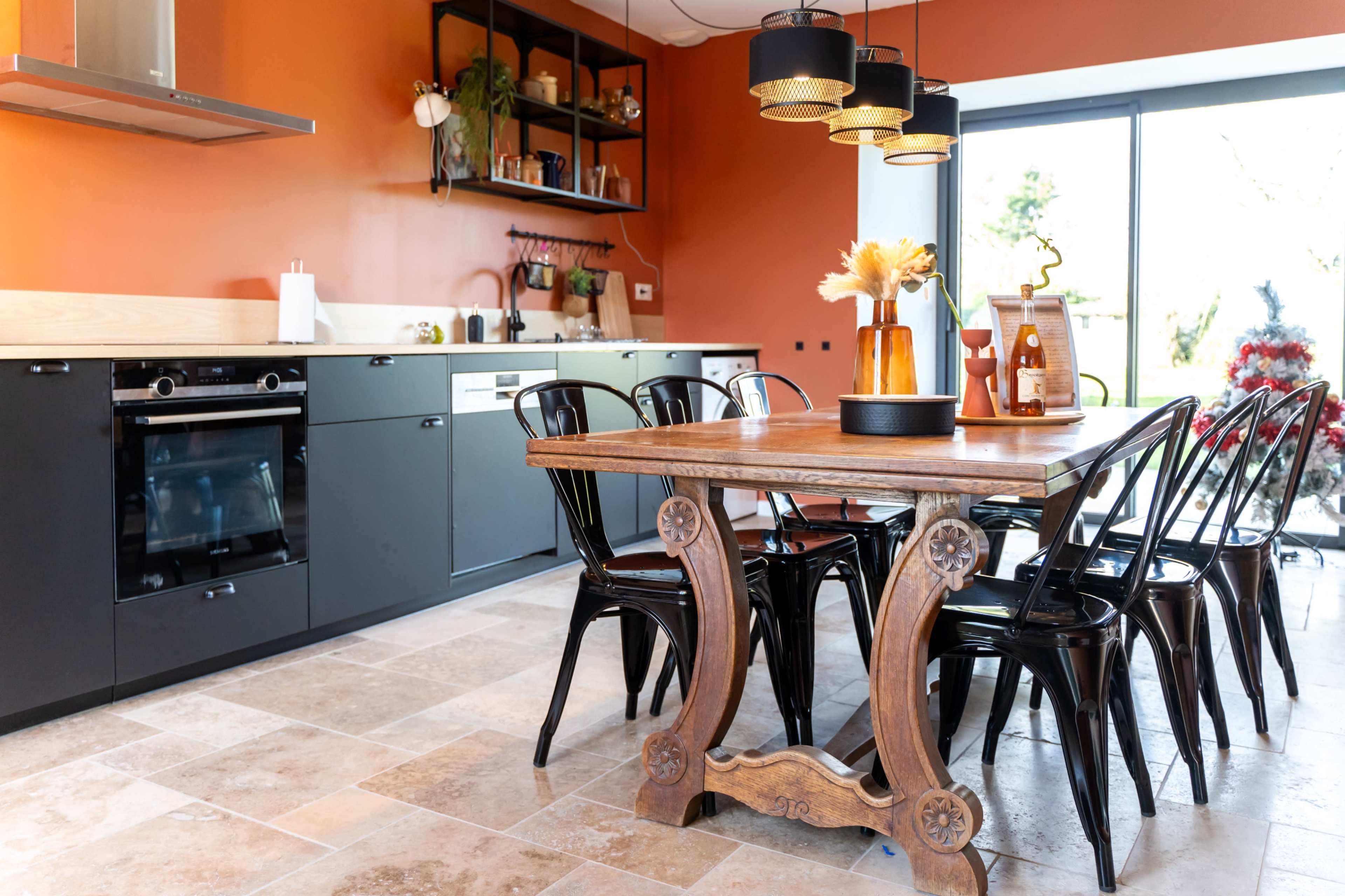 The image shows a modern kitchen with an orange accent wall, featuring a wooden dining table surrounded by black chairs, and sleek kitchen appliances.