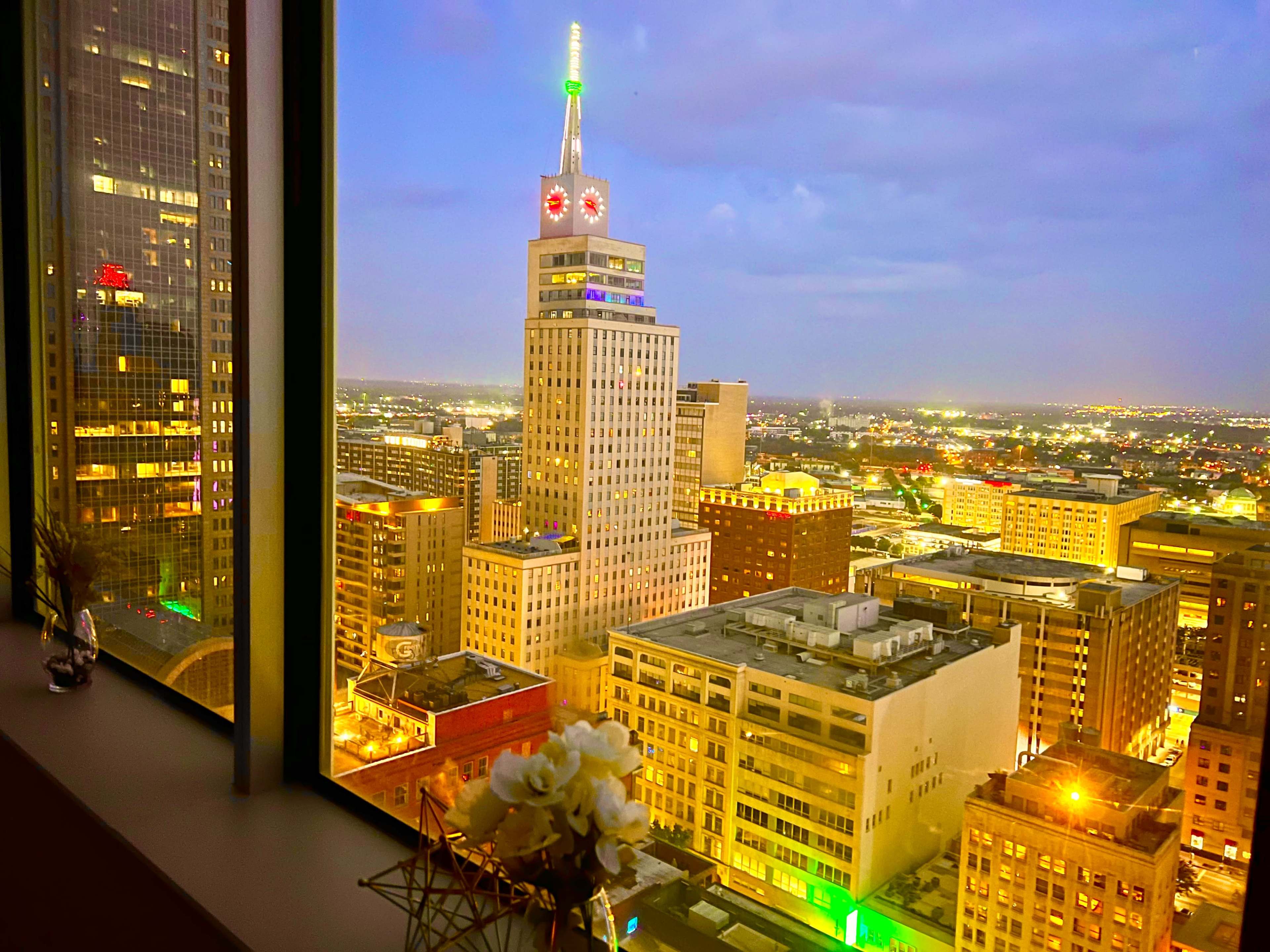 A city skyline at dusk, featuring a tall building with a brightly lit top and various illuminated structures below.