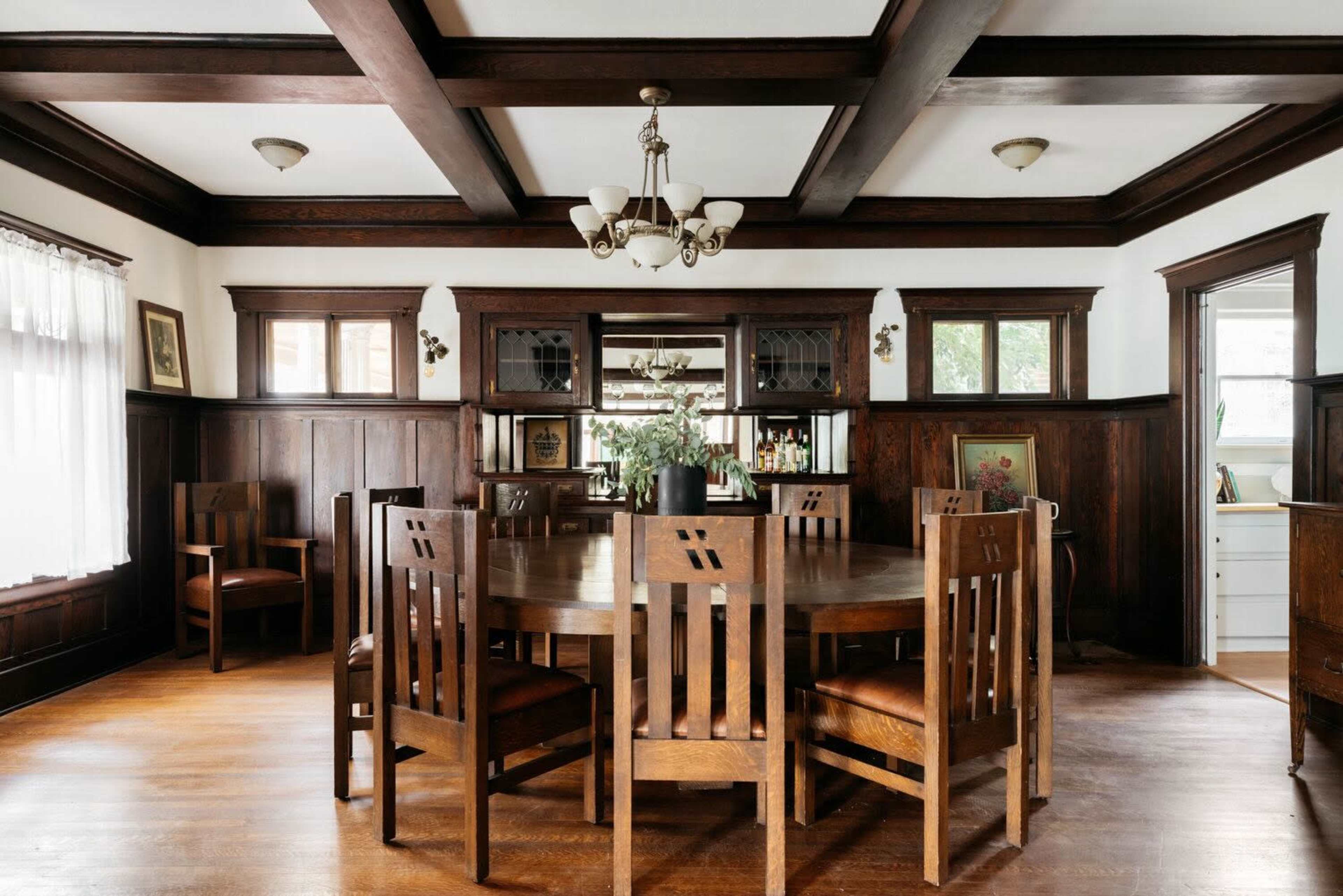 The image shows a wooden dining room with a large circular table surrounded by eight chairs, framed by dark wood paneling and a built-in cabinet.