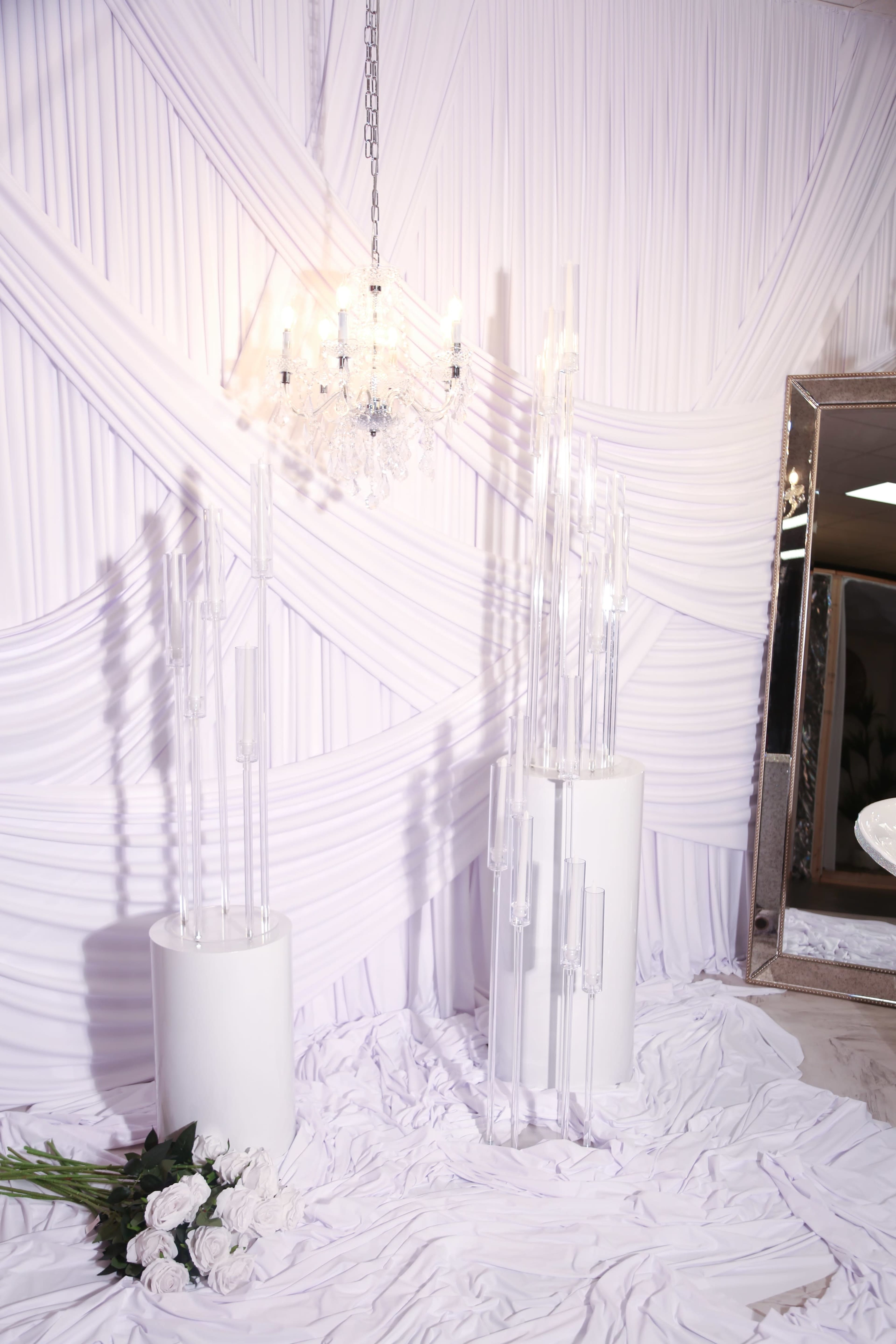 The scene features two tall, clear decorative columns on white pedestals, set against a backdrop of draped white fabric and a crystal chandelier.