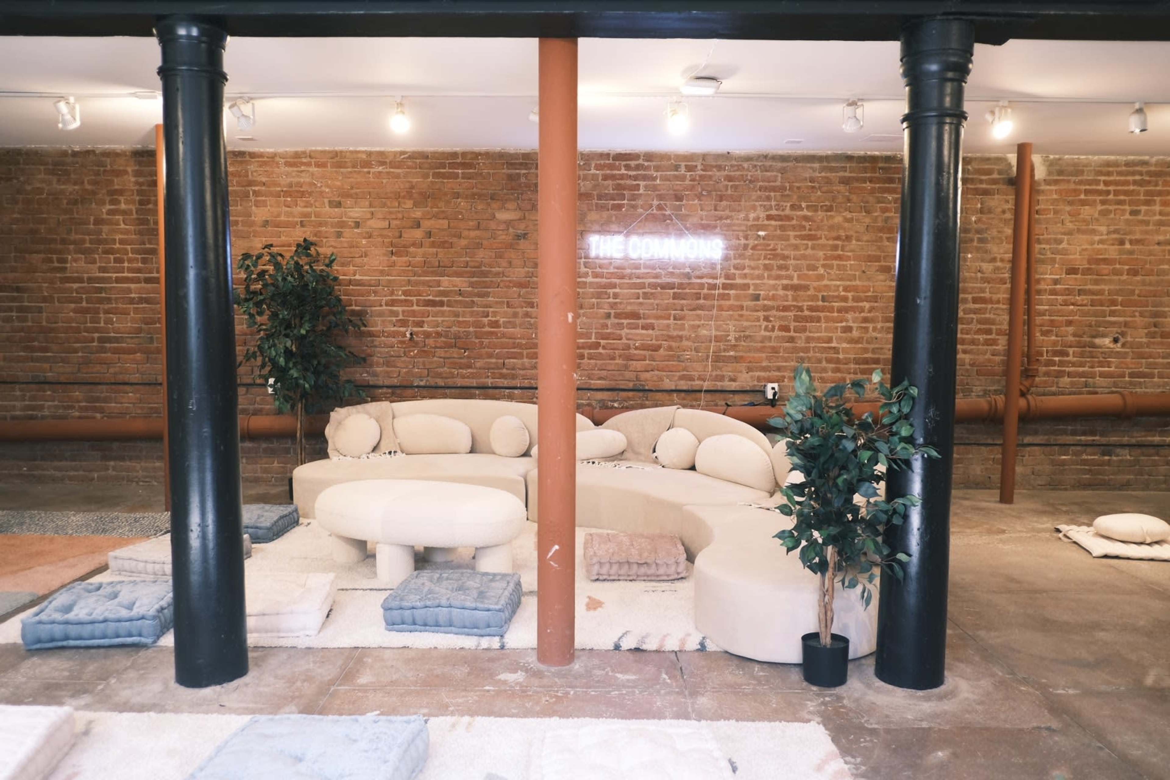 A cozy communal lounge area features a curved sofa, a round table, and soft floor cushions surrounded by brick walls and illuminated by overhead lights.