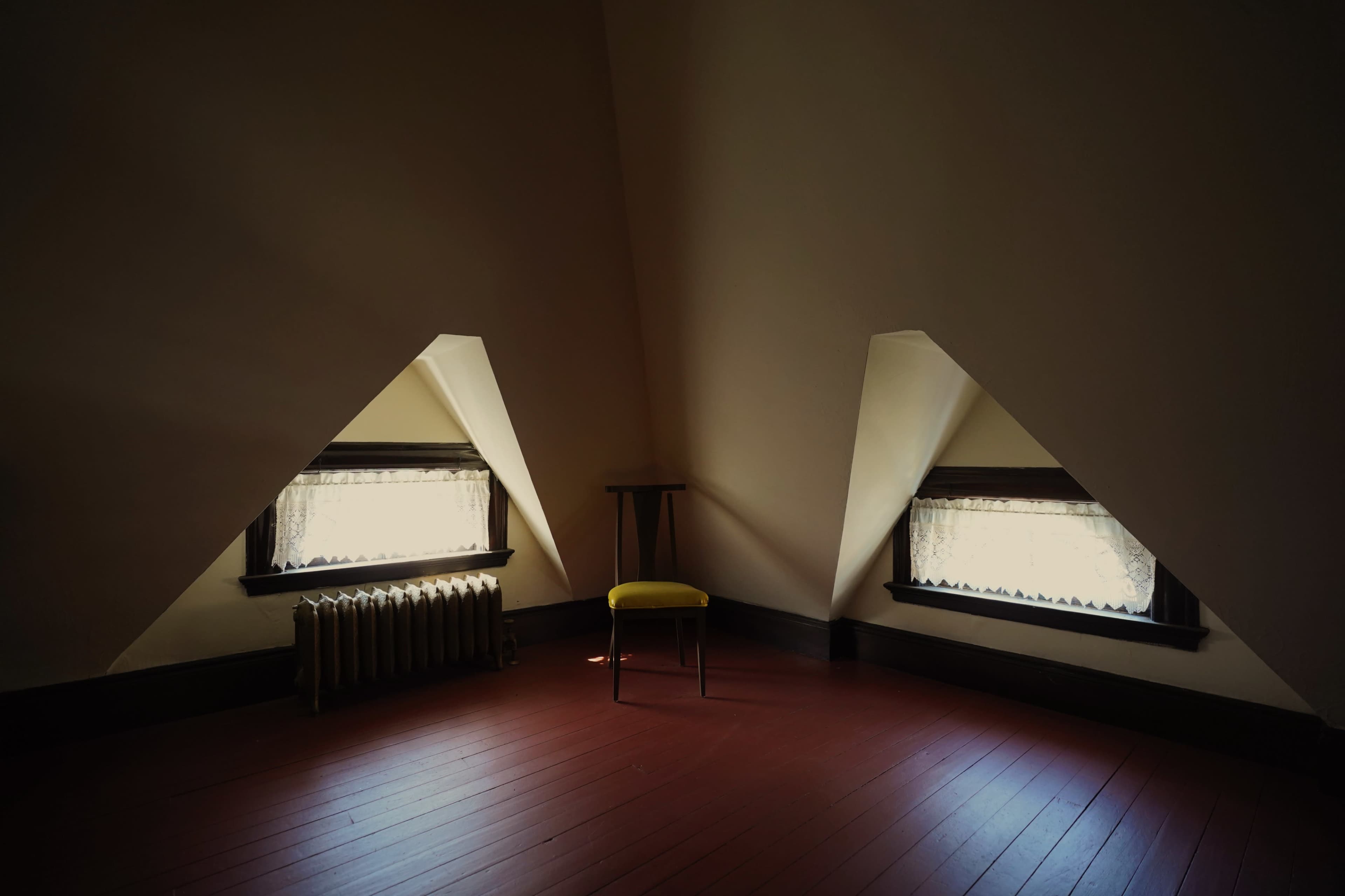 A small, sparsely furnished attic room features two triangular windows, a wooden chair, and a radiator along the wall.
