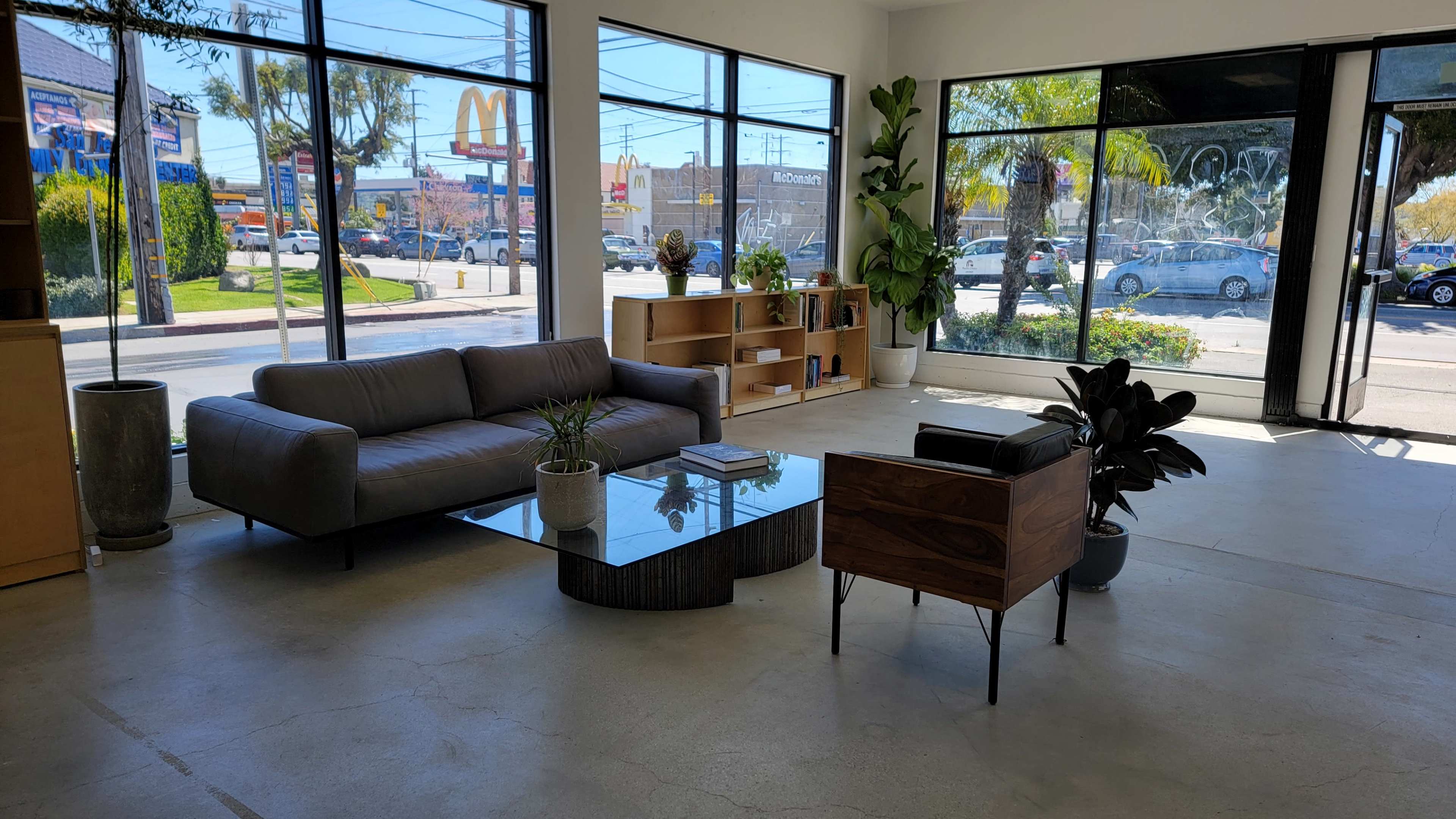 The interior of a modern, sparsely furnished room features a gray sofa, a glass coffee table, and a wooden chair, with large windows displaying a busy street outside.