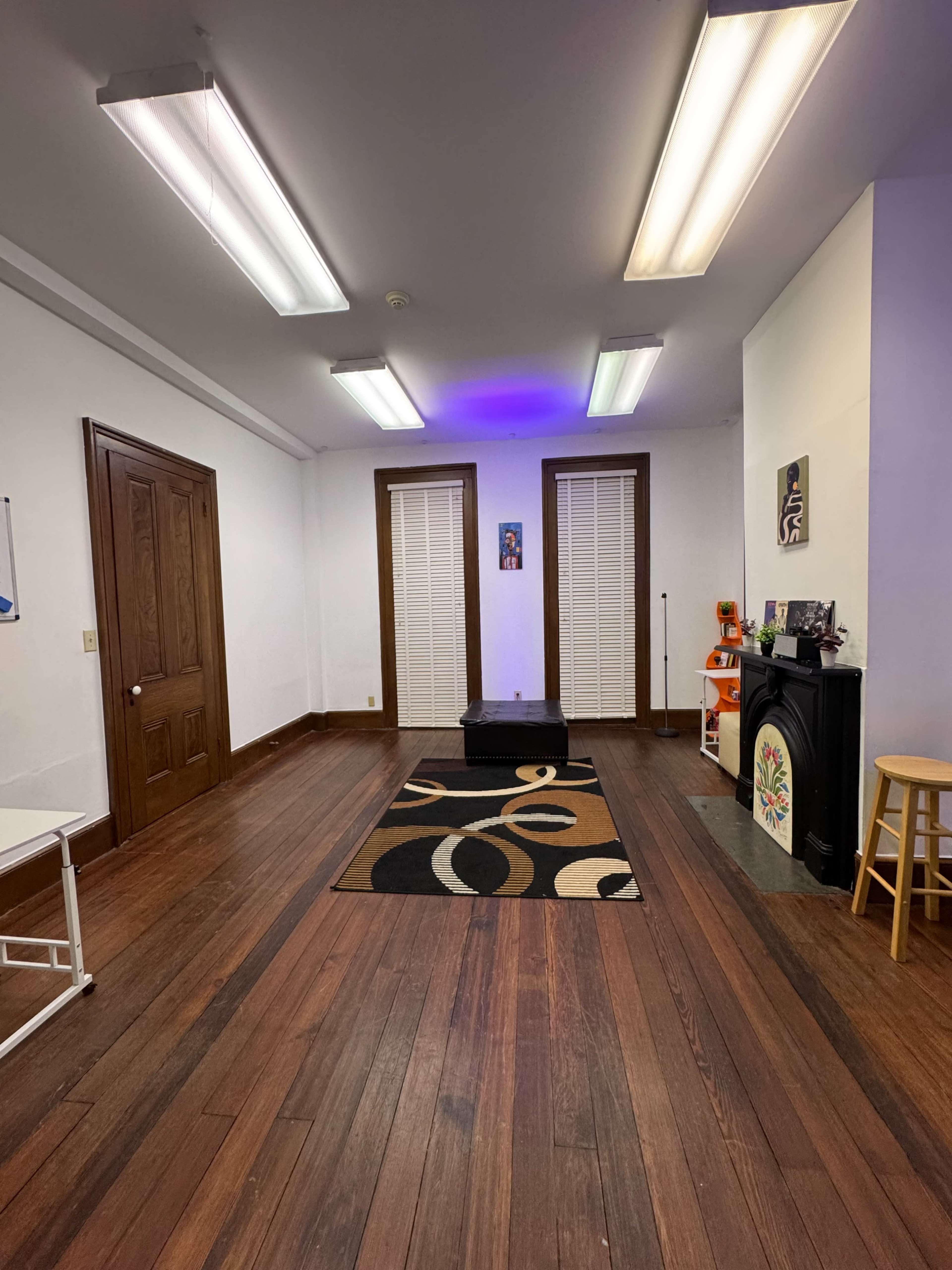 The image shows an empty room with wooden flooring, white walls, two windows with blinds, and a patterned rug in the center.