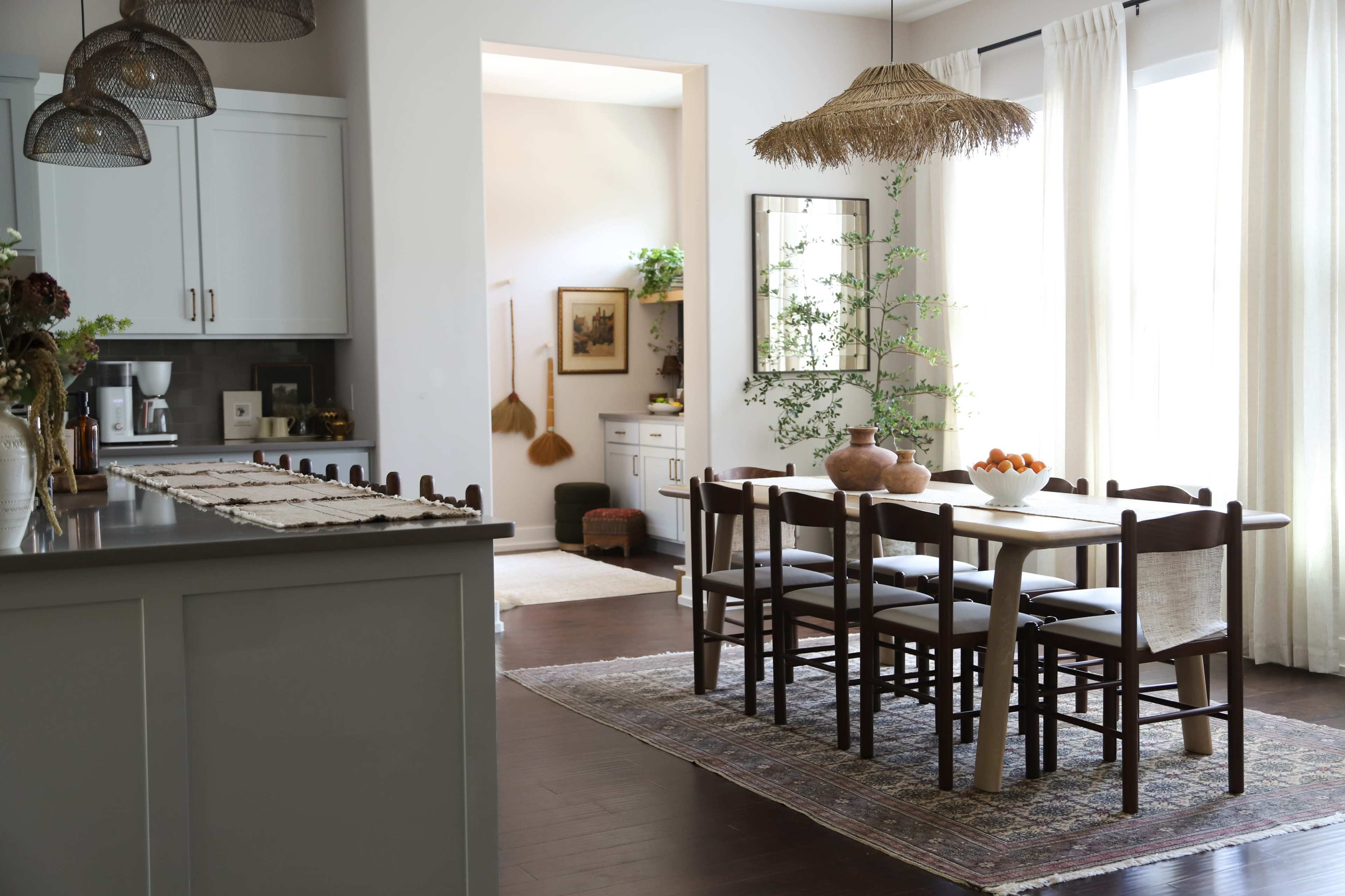 A modern kitchen and dining area feature a wooden dining table surrounded by six chairs, with natural light streaming in through large windows and a hanging light fixture made of woven material.
