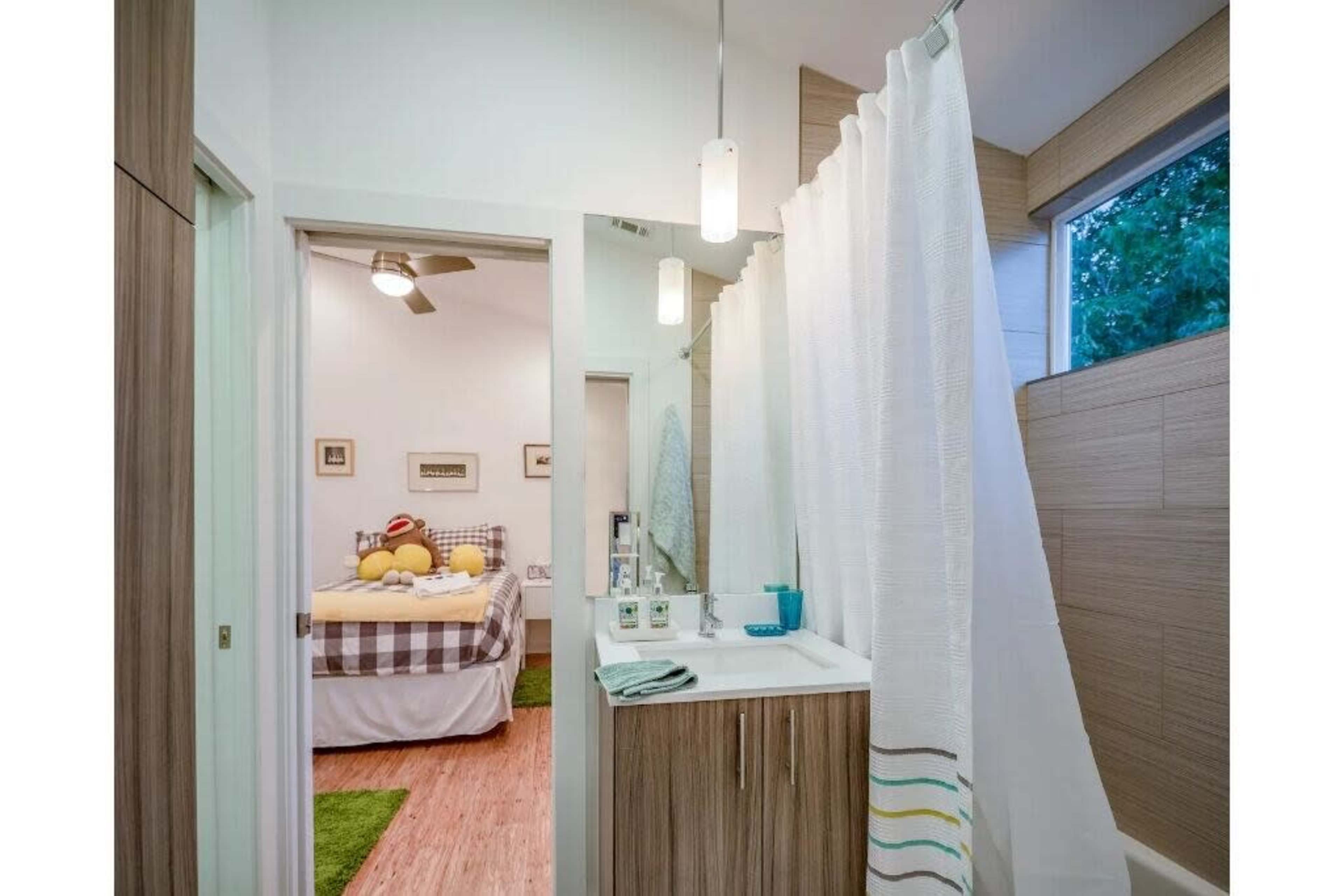 A bathroom features a white shower curtain, modern fixtures, and a door leading to a bedroom with a checkered bedspread and a few decorative pillows.