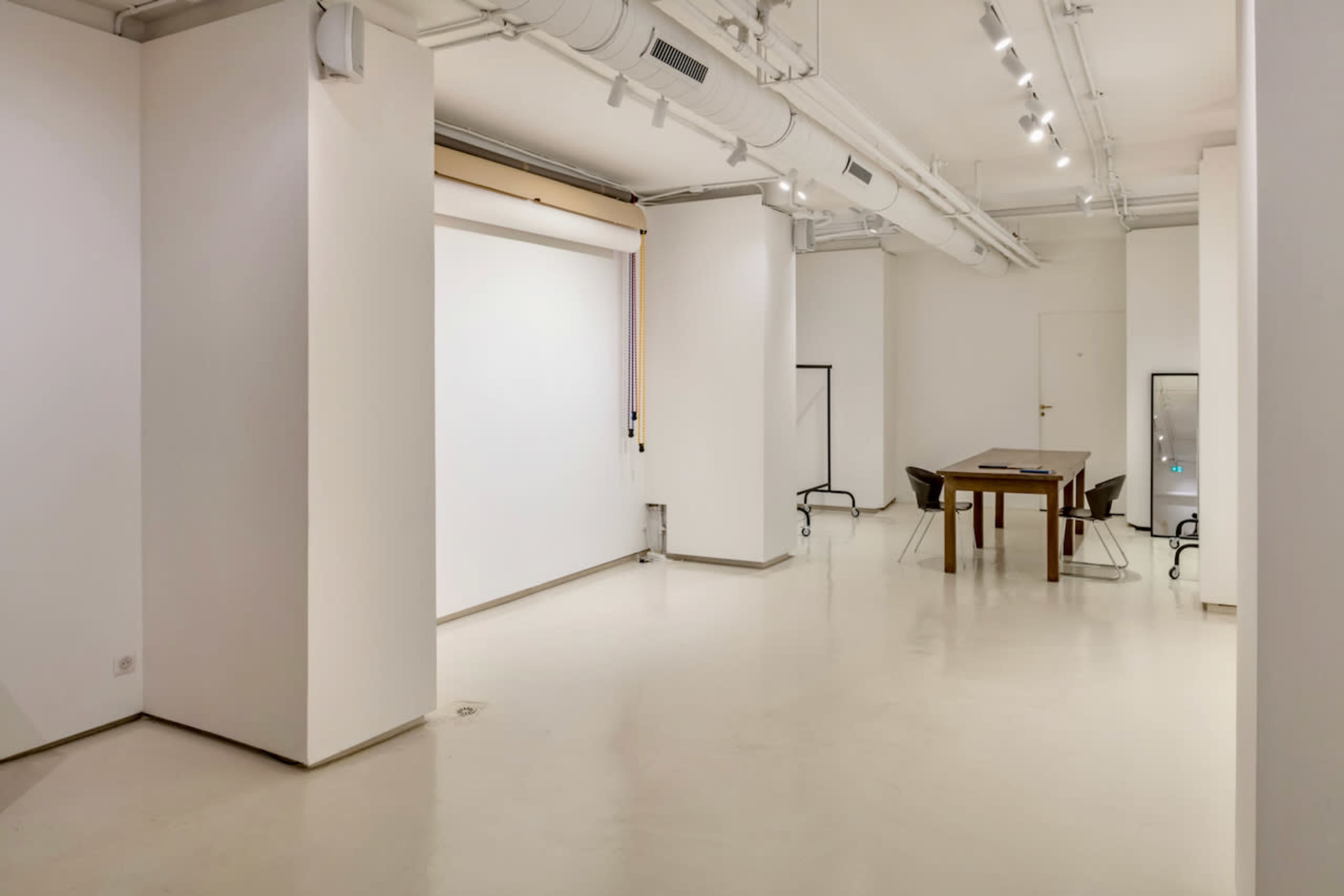 The image shows a spacious, well-lit room with white walls, a large empty wall area, and a wooden table surrounded by chairs.