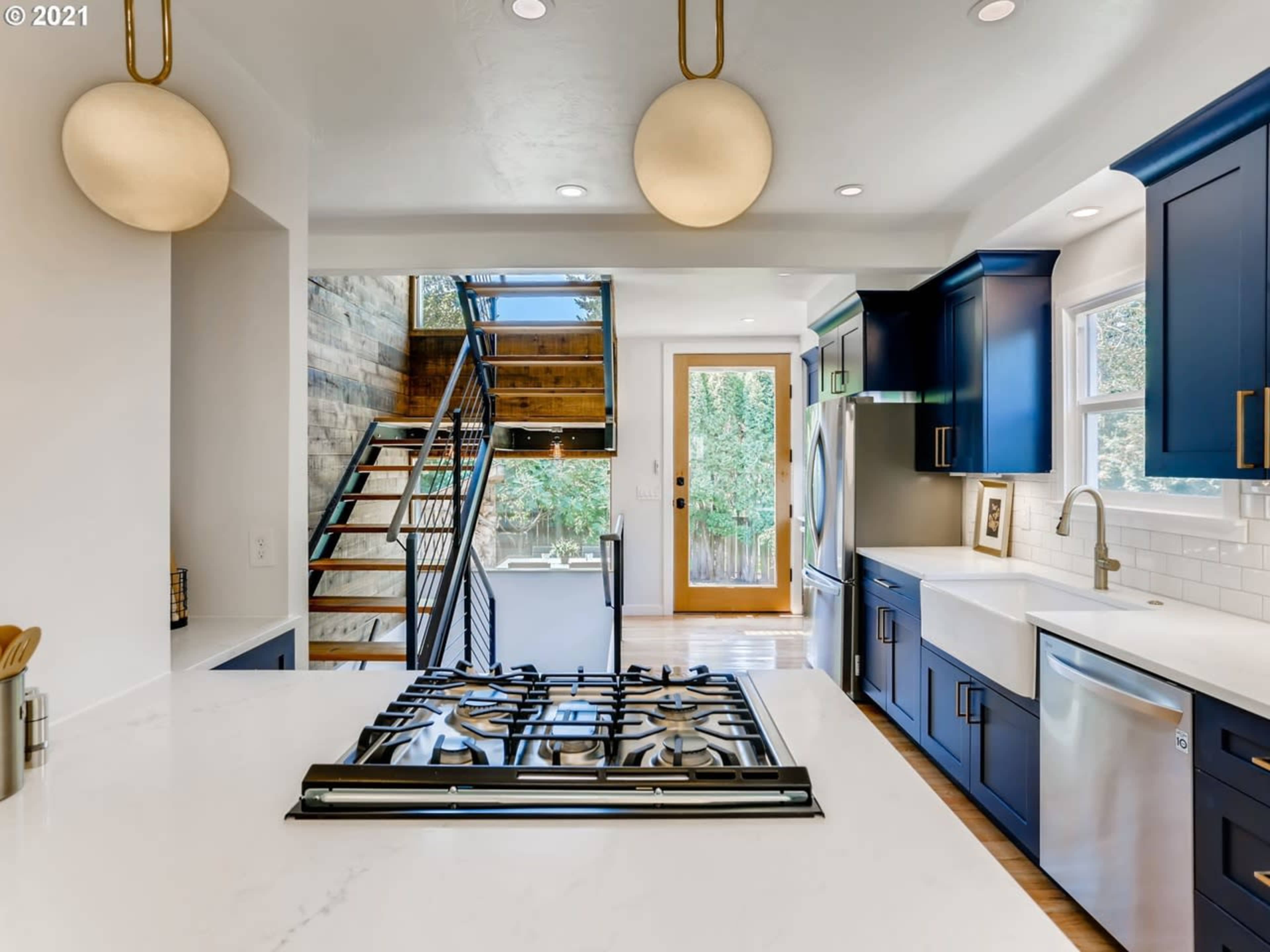 The image shows a modern kitchen with blue cabinetry, a white countertop, and a view of a staircase leading to an upper level.