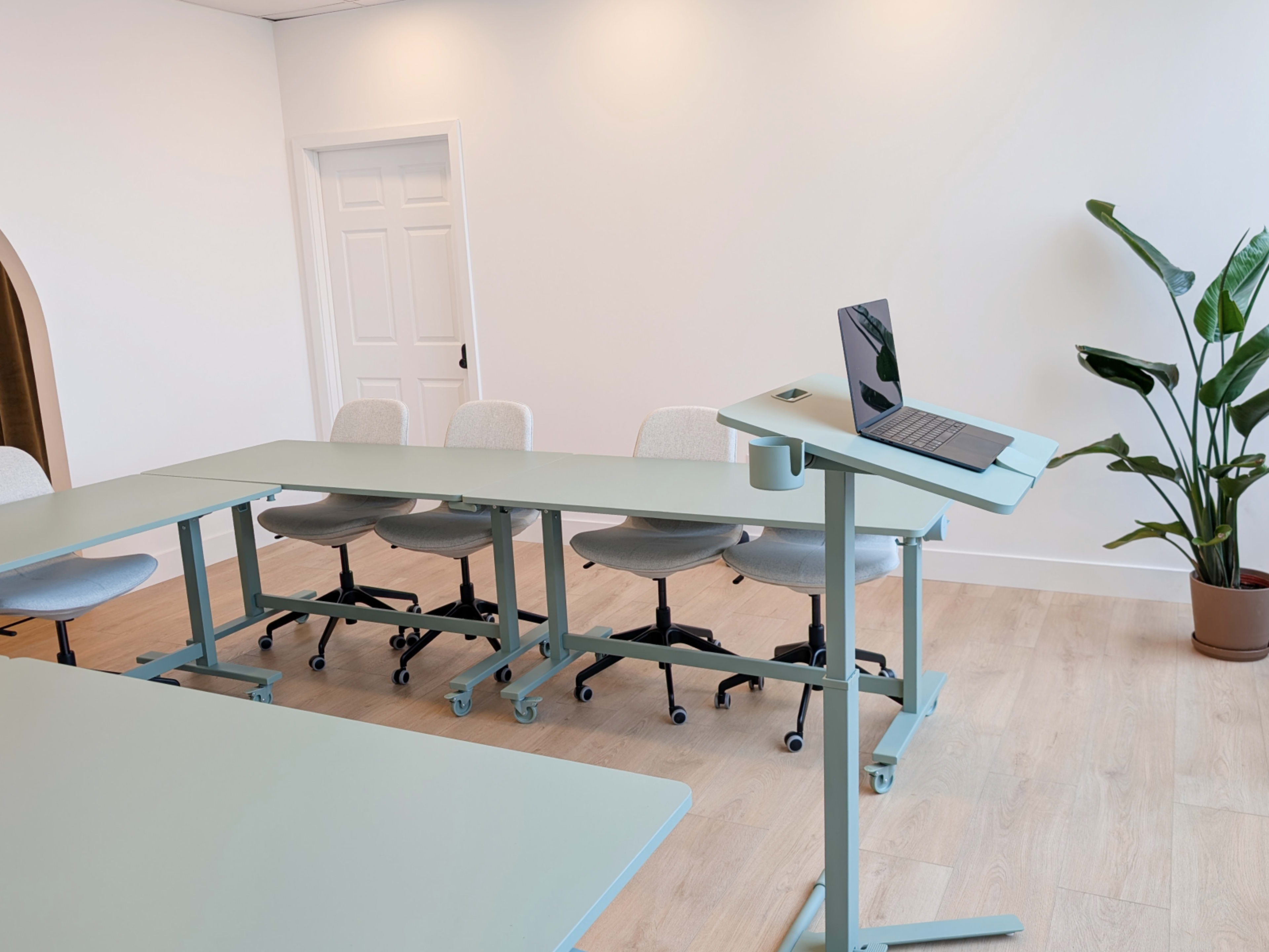 The image shows a modern, minimalistic meeting room with a light green table, chairs on wheels, and a laptop on a stand beside a potted plant.