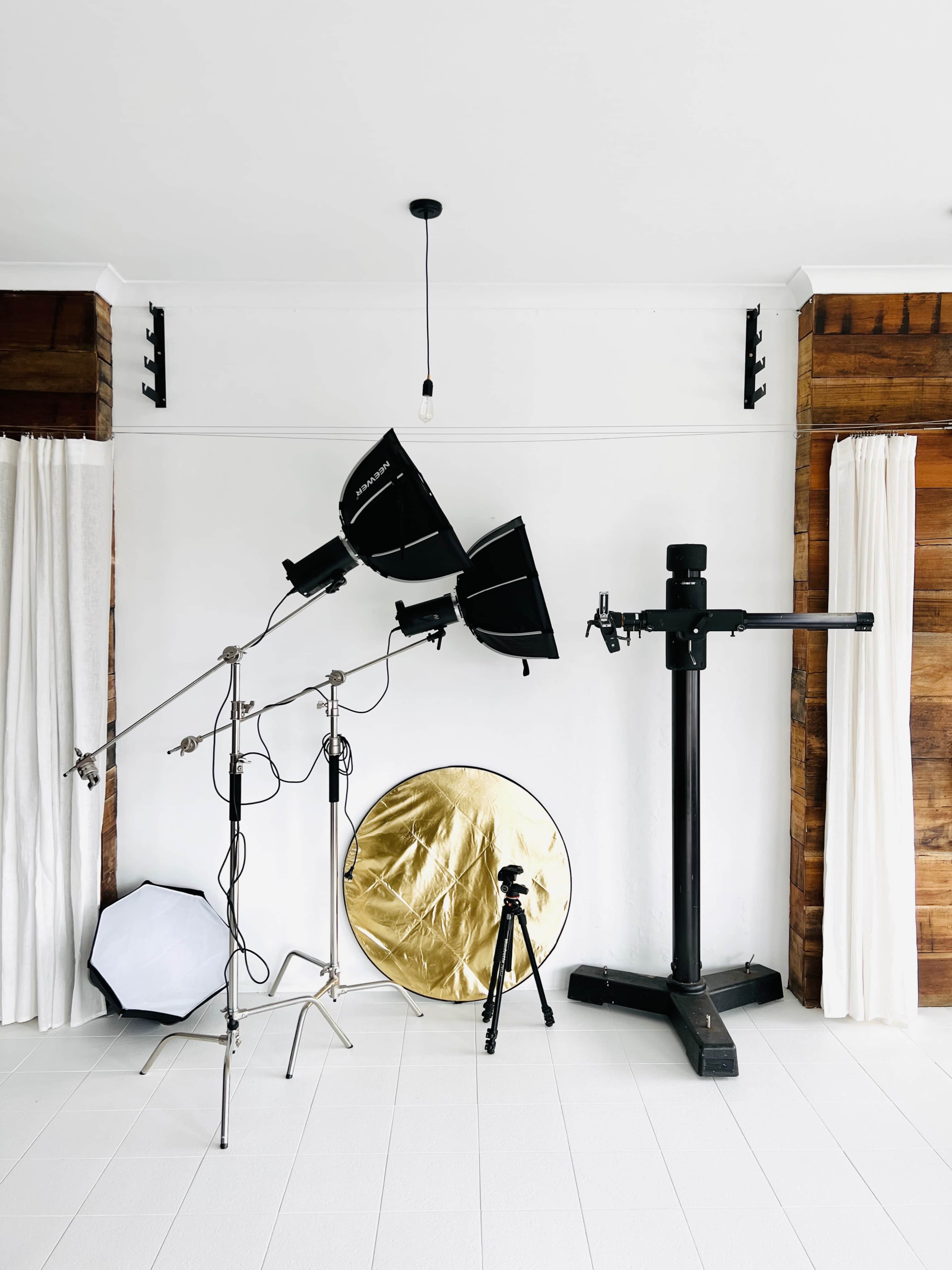 The image depicts a photography studio setup with two light stands, a gold reflector, and a tripod against a white wall.